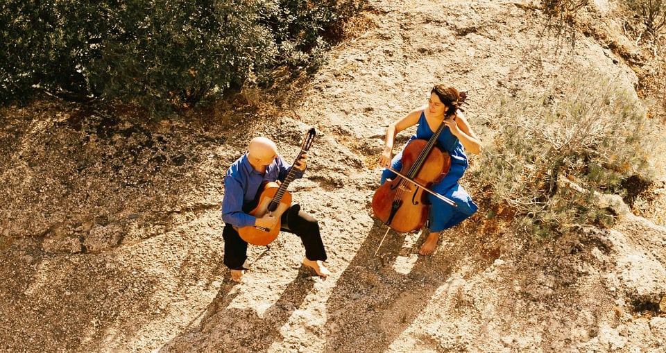 Zwei Musiker der Passion Azul in blauer Kleidung mit Instrumenten, von oben fotografiert, stehen auf steinigem Boden mit Büschen