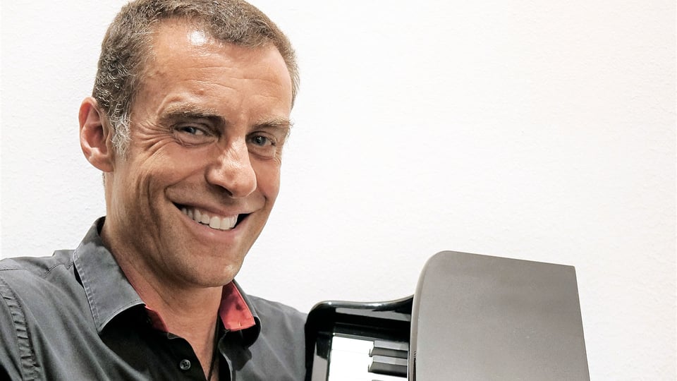 Martin Schmitt im dunklen Hemd in Nahaufnahme, seitlich zur Kamera blickend und lächelnd, neben einem kleinen Tasteninstrument vor hellem Hintergrund