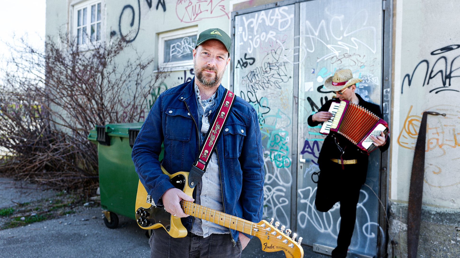 Der Künstler mit Gitarre vor einer Hauswand mit Graffiti, an der ein weiterer Musiker lehnt