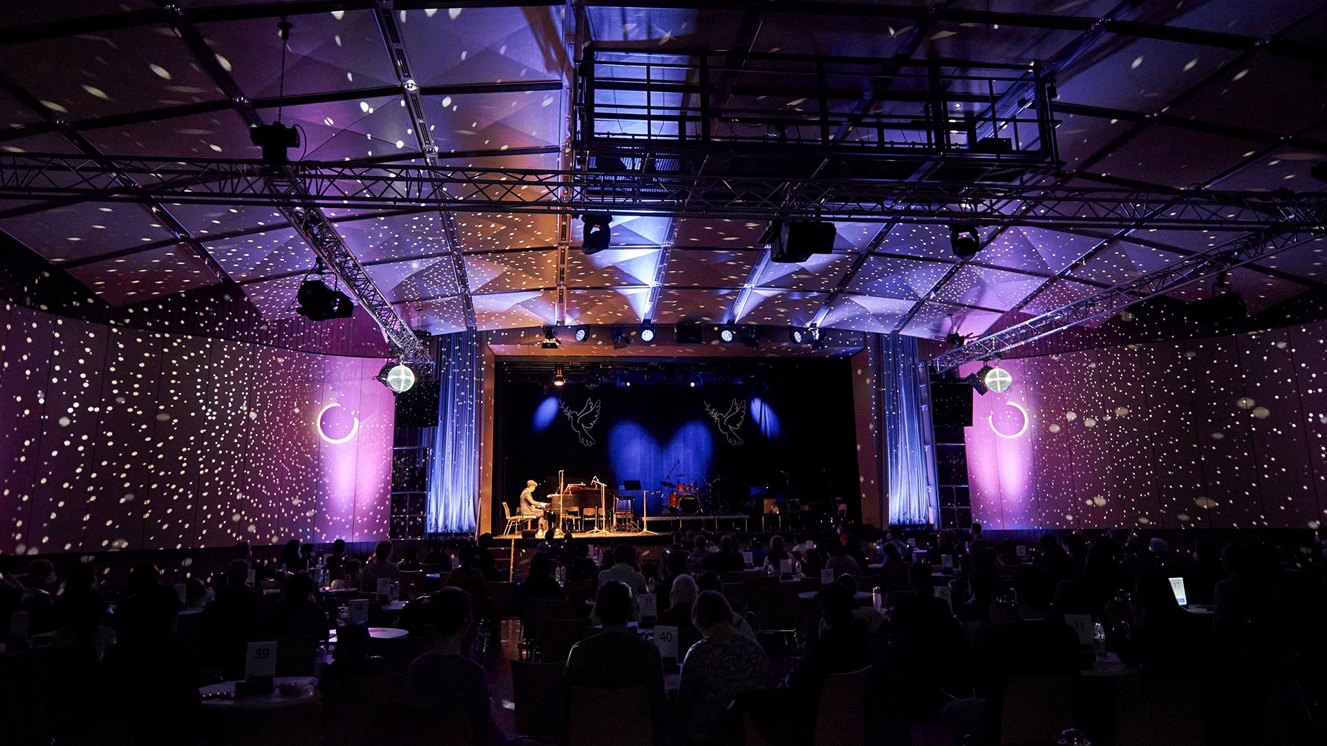 Konzertsaal mit großem sitzendem Publikum vor einer Bühne. Die Halle ist mit violett blauem Licht und zahlreichen punktförmigen Lichtprojektionen beleuchtet, während auf der Bühne ein Flügel und weitere Instrumente aufgebaut sind.