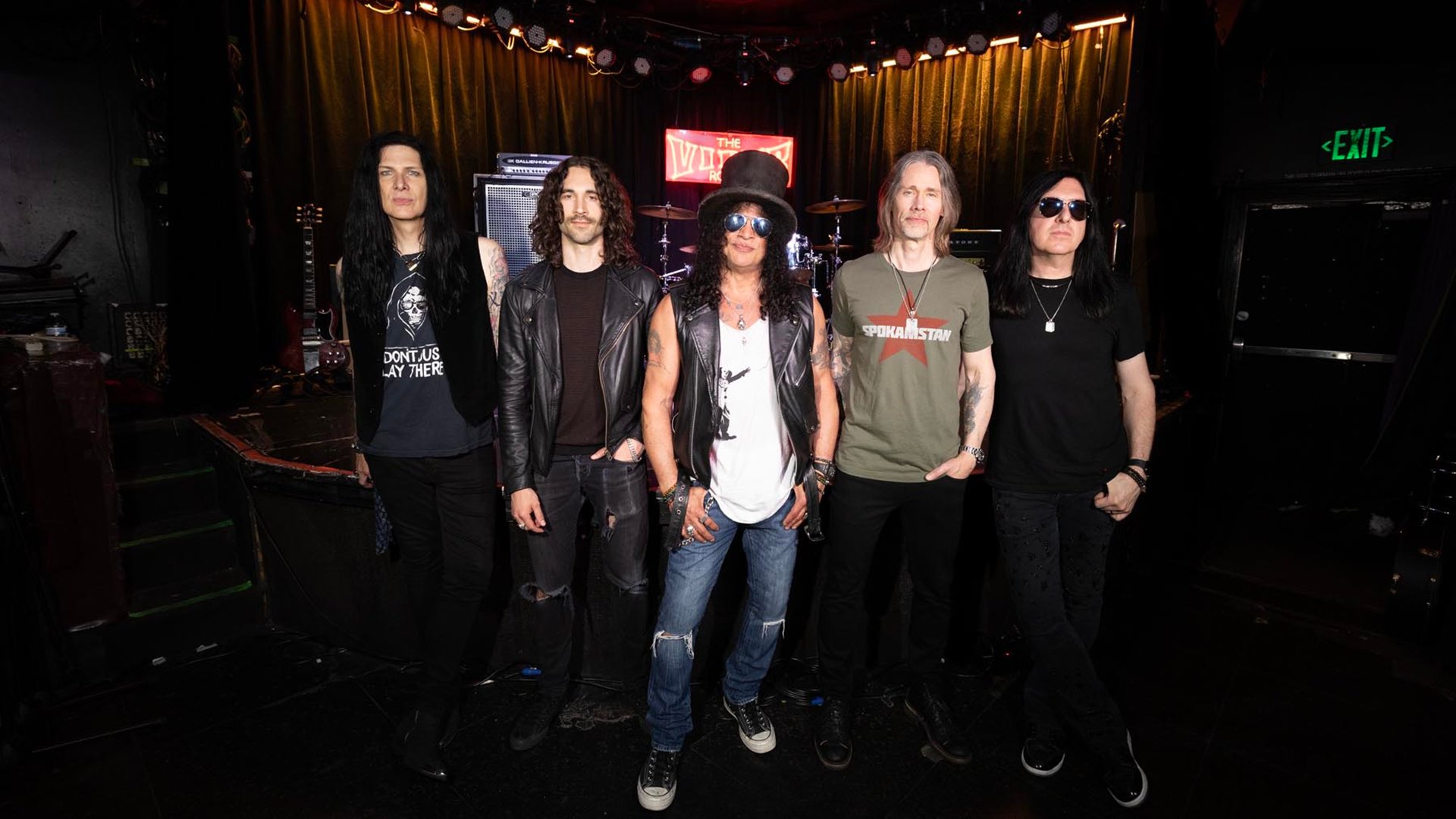 Slash und Myles Kennedy mit drei Personen der Band Conspirators blicken in die Kamera. Im Hintergrund eine Bühne mit Schlagzeug und Gitarre.