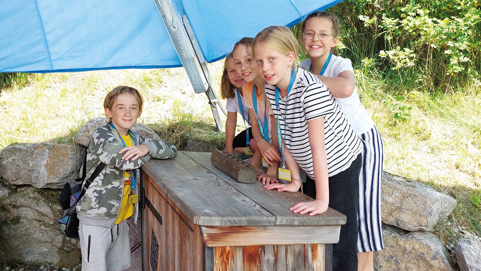 Fünf Kinder stehen gemeinsam an einer Holzkiste im Freien unter einem Sonnenschutz, tragen Namensschilder und befinden sich in einer grünen Umgebung mit Bäumen und Steinen