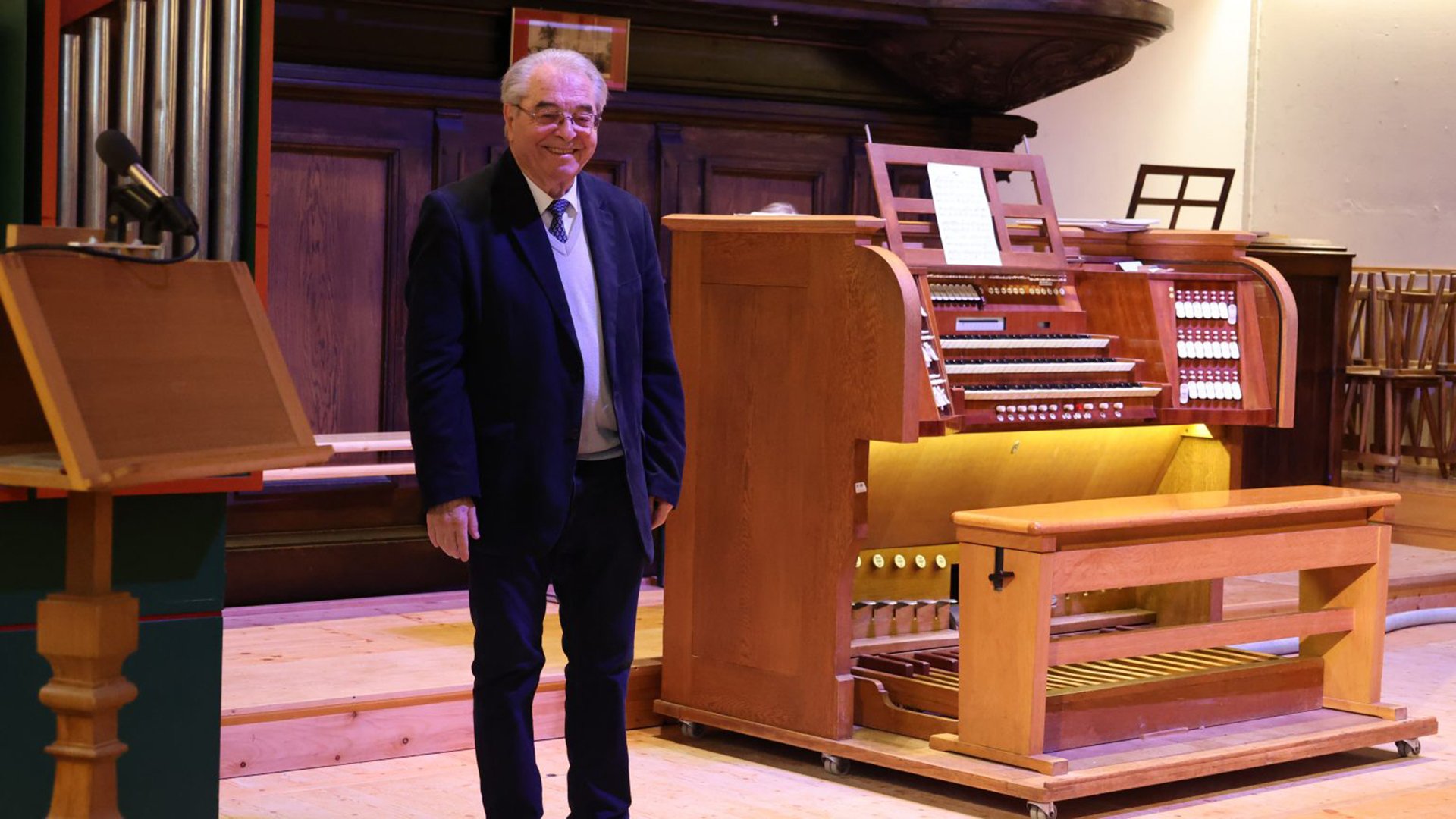 Person steht neben einer Orgel in einem holzverkleideten Konzertsaal, mit Notenständern und Sitzgelegenheiten im Hintergrund.