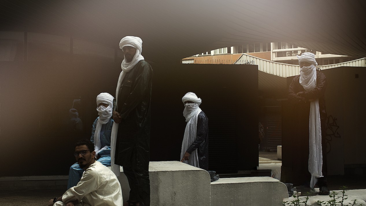 Vier vermummte Gestalten mit weißen Turbanen, ein weiterer Mann ohne, stehem hinter Betonklötzen