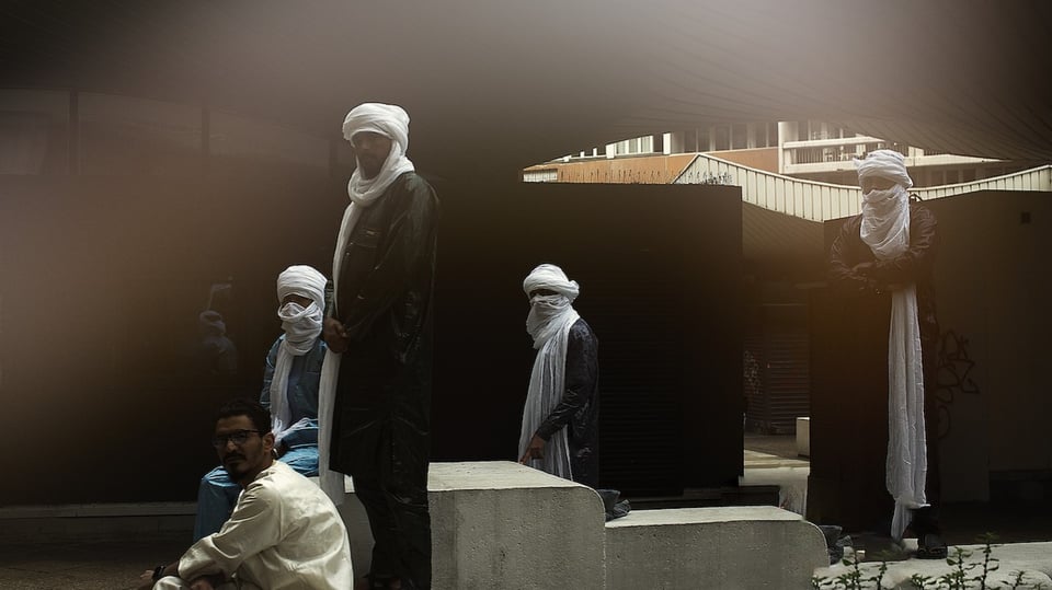 Vier vermummte Gestalten mit weißen Turbanen, ein weiterer Mann ohne, stehem hinter Betonklötzen