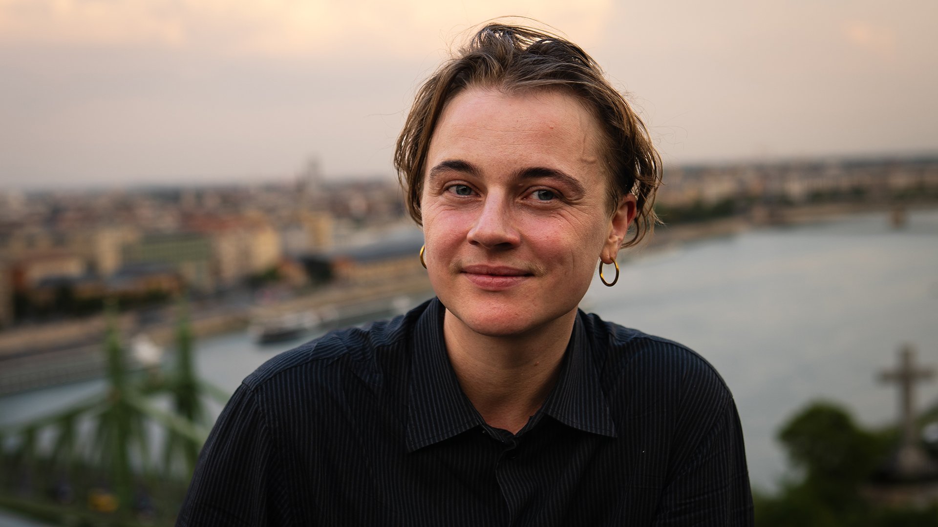 Darida Muri in schwarzem Hemd mit goldfarbenen Creolen, vor unscharfer Stadt- und Flusslandschaft bei Abendlicht
