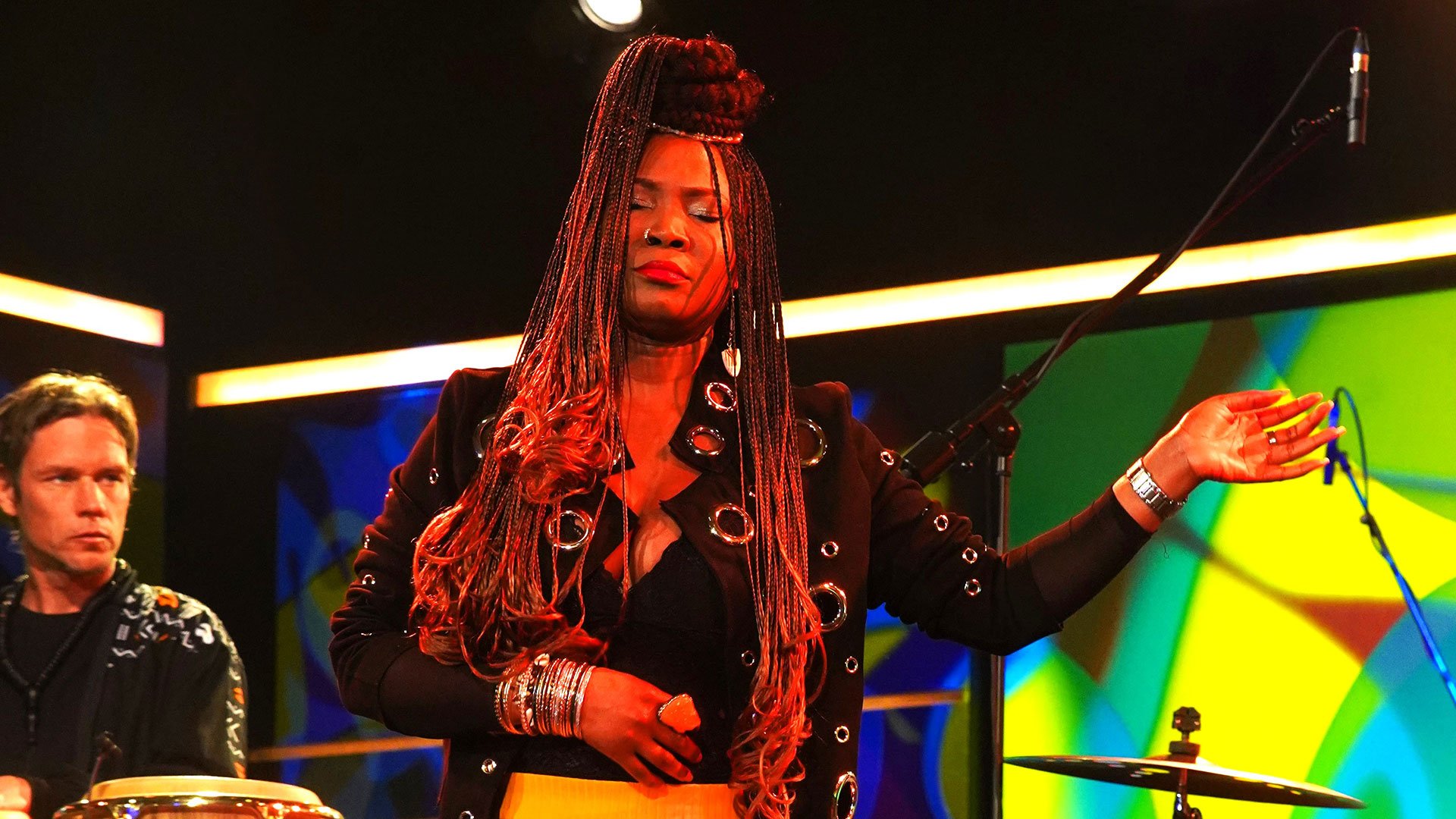 Foto für die Veranstaltung JUSTINA LEE BROWN mit der Künstlerin auf einer Bühne, im Hintergrund ein Musiker und farbige Visuals.