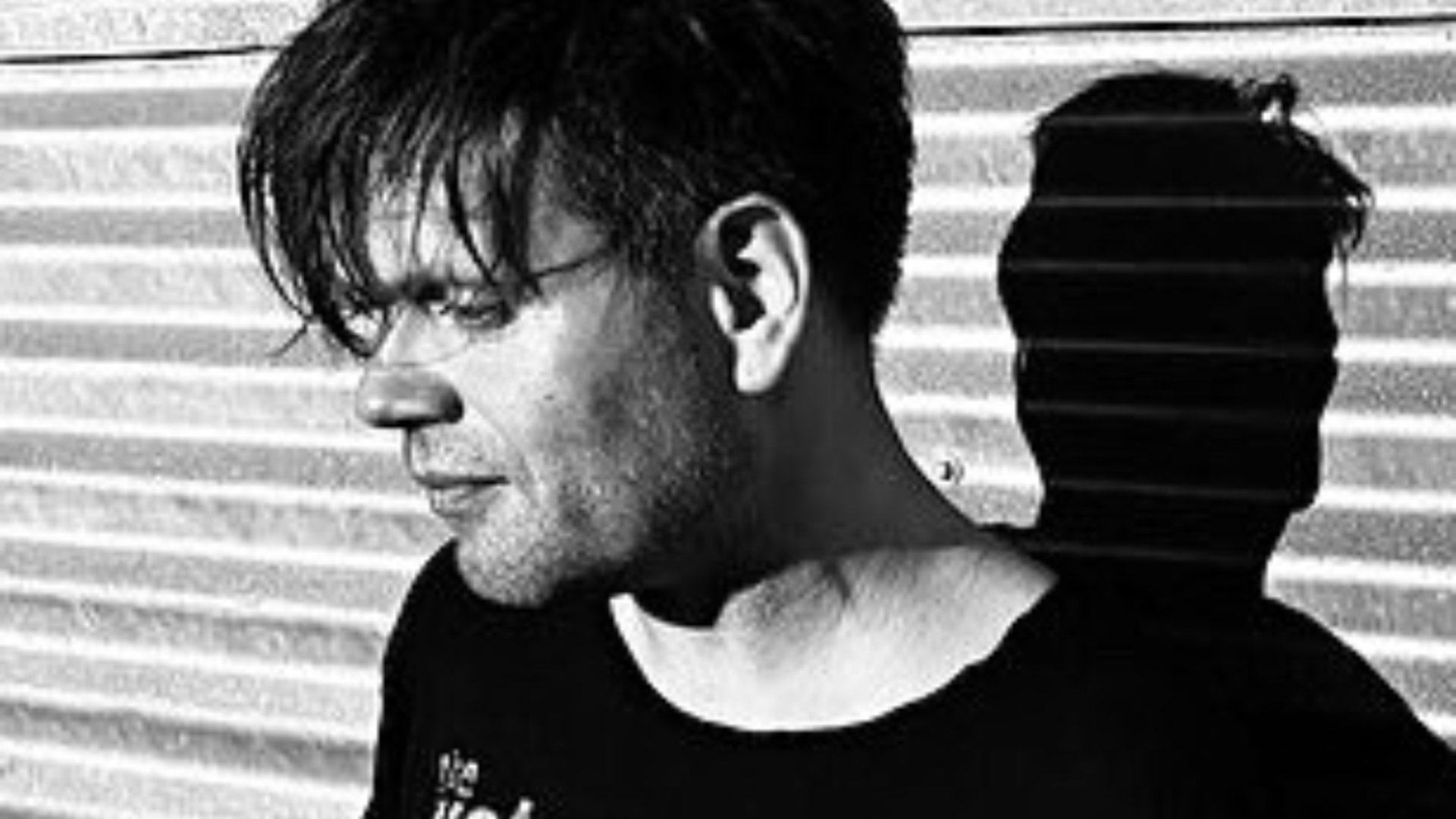 Schwarz‑weißes Porträtmotiv zu Trentemøller: seitliche Ansicht einer Person mit dunklem T‑Shirt vor geriffelter Metallwand, starkes Licht erzeugt einen deutlichen Schlagschatten im Hintergrund; reduzierter, kontrastreicher Look für ein elektronisches Live‑Konzert.