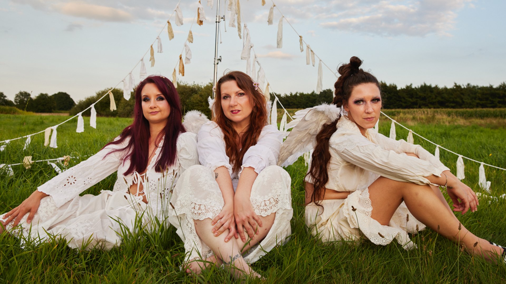 Drei Damen in weißen Kleidern sitzen auf einer Wiese, über ihnen weiße WImpel