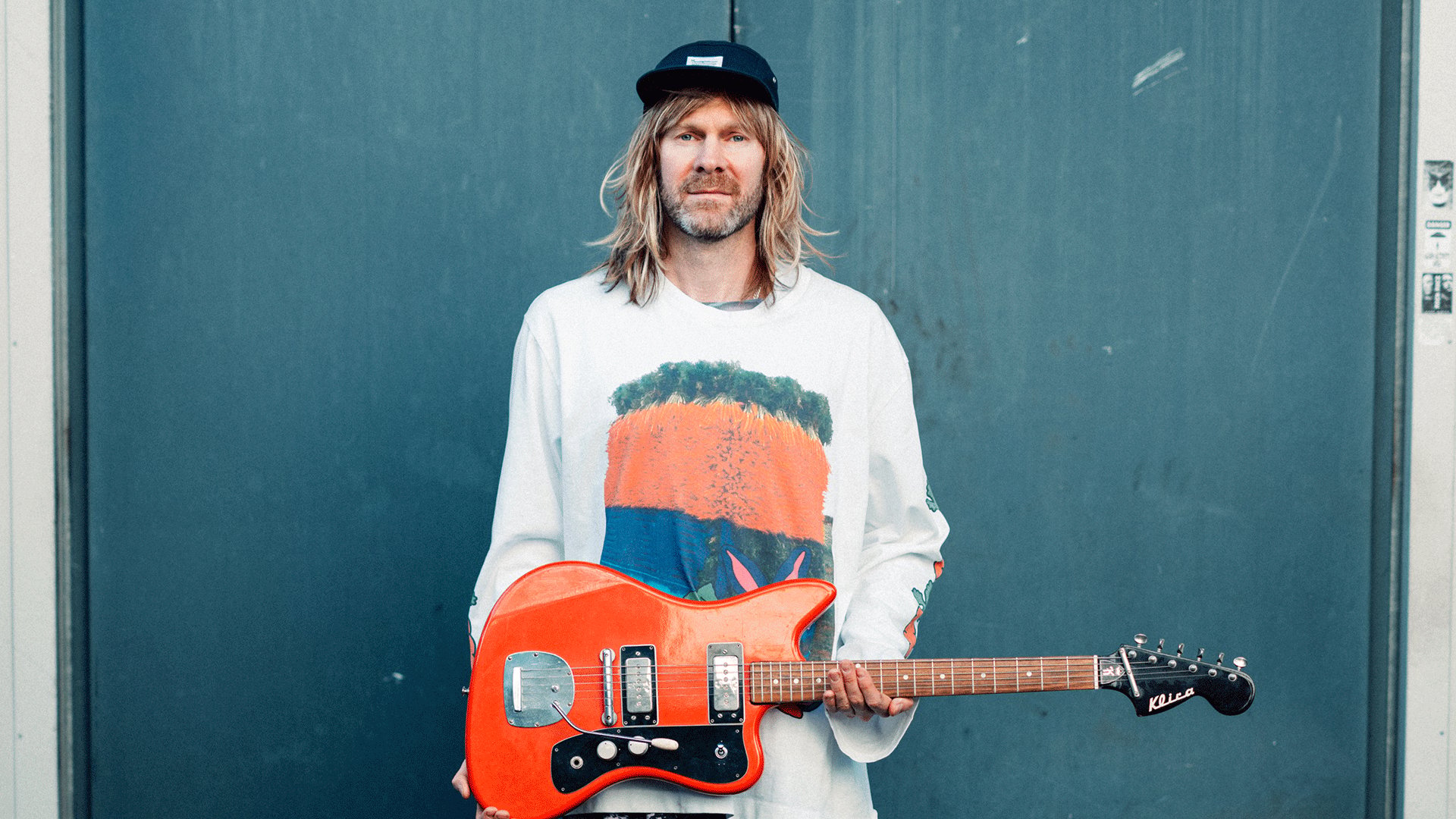 Porträt einer Person mit langem blondem Haar und schwarzer Kappe. Die Person hält eine orange E Gitarre vor dem Körper und trägt ein helles Oberteil mit großflächigem Motiv.