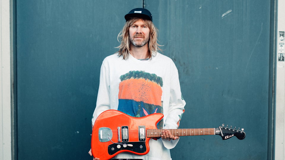 Porträt einer Person mit langem blondem Haar und schwarzer Kappe. Die Person hält eine orange E Gitarre vor dem Körper und trägt ein helles Oberteil mit großflächigem Motiv.