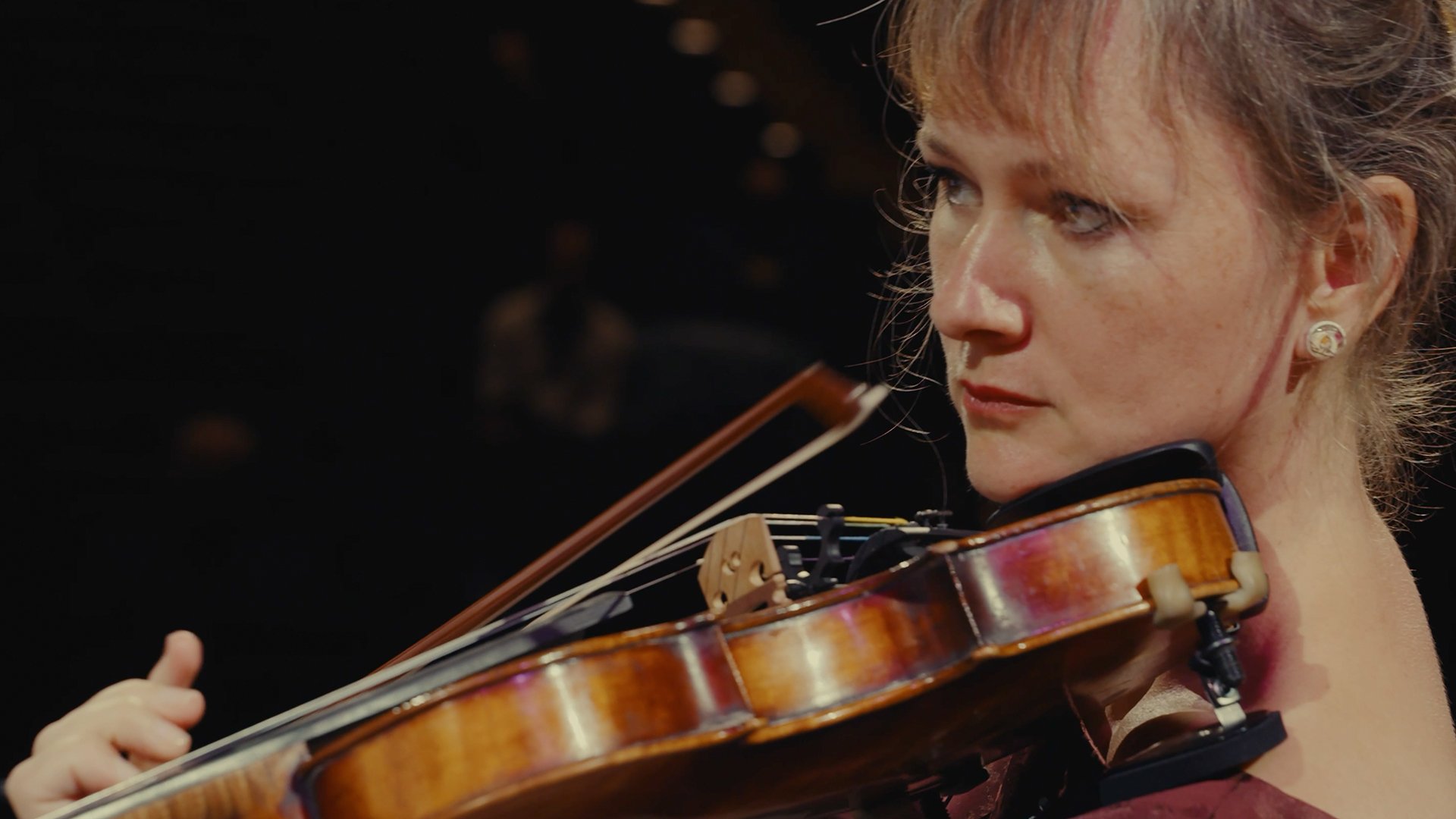 Detailreiche Bühnenaufnahme einer Person beim Violinespiel: Die Violine liegt auf der Schulter, der Bogen wird dicht an den Saiten geführt. Warmes Bühnenlicht hebt das Holz des Instruments hervor, dunkler Hintergrund mit unscharfen Lichtpunkten. Ruhiges, konzentriertes Motiv für eine klassische / neoklassische Live‑Performance mit Streichinstrument.