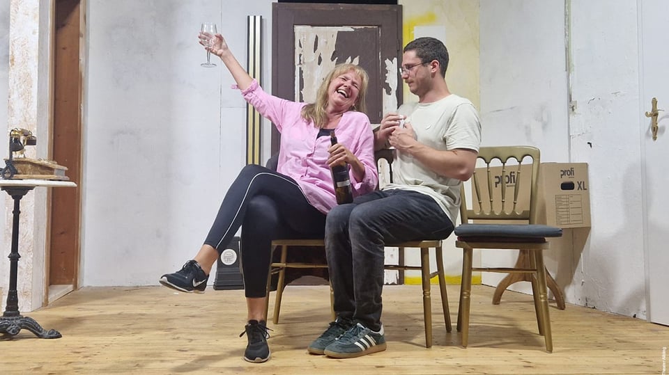 Theaterszene mit zwei sitzenden Personen auf einer Bühne vor einer Wohnungs-Kulisse.