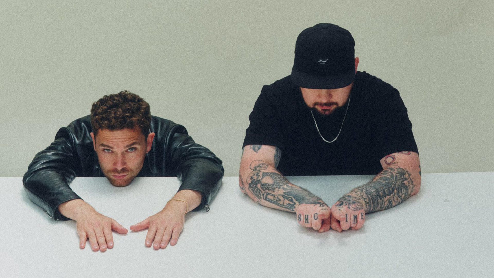 Bandfoto von Royal Blood mit zwei Bandmitgliedern an einem weißen Tisch vor hellem Hintergrund, eine Person in schwarzer Lederjacke mit dem Kopf auf dem Tisch und direktem Blick zur Kamera, die andere mit schwarzer Kappe und tätowierten Unterarmen, die Hände vor sich auf dem Tisch verschränkt.