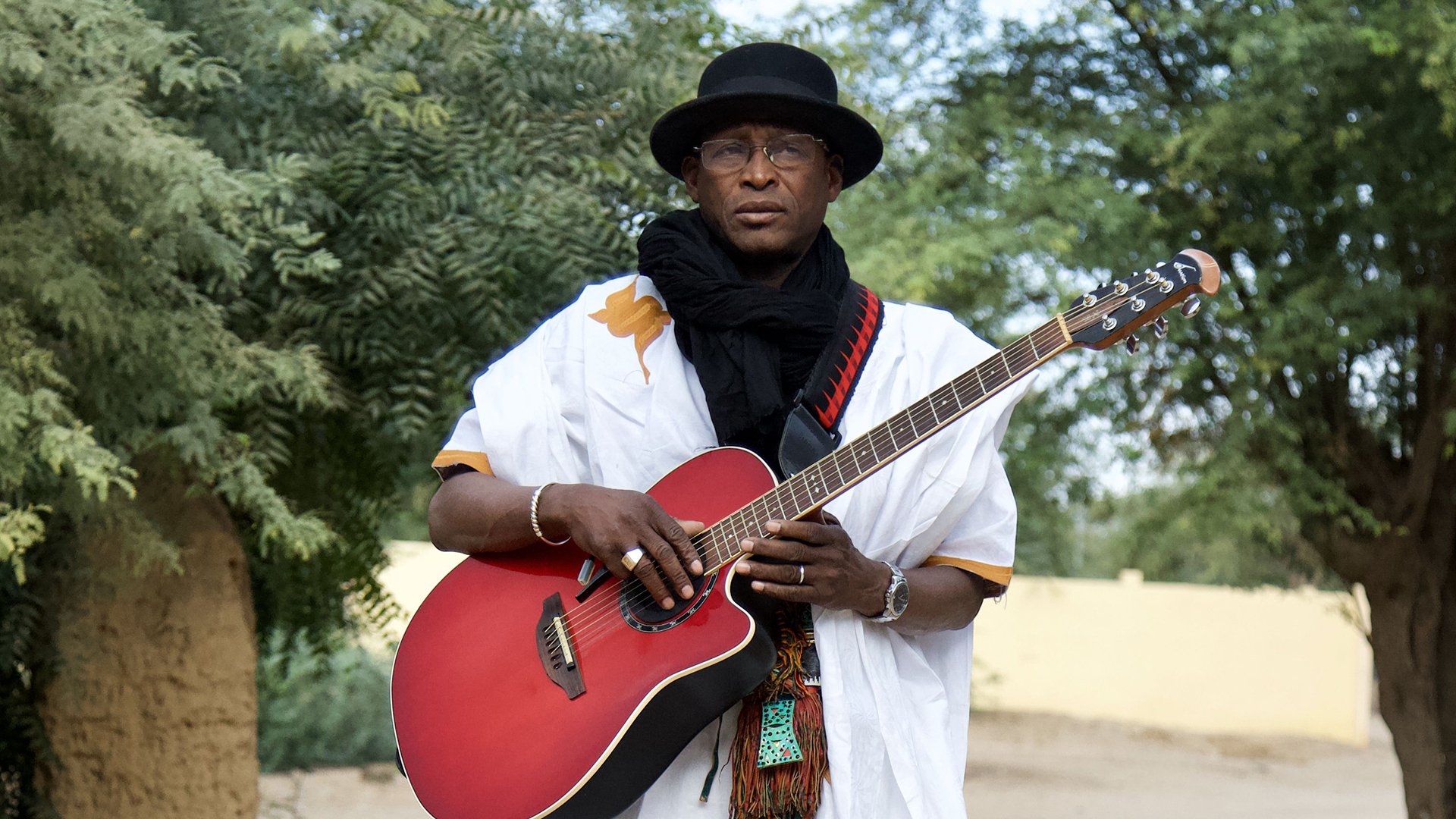 Foto des Künstlers Afel Bocoum mit einer roten Gitarre in den Händen, in der Natur zwischen Bäumen.
