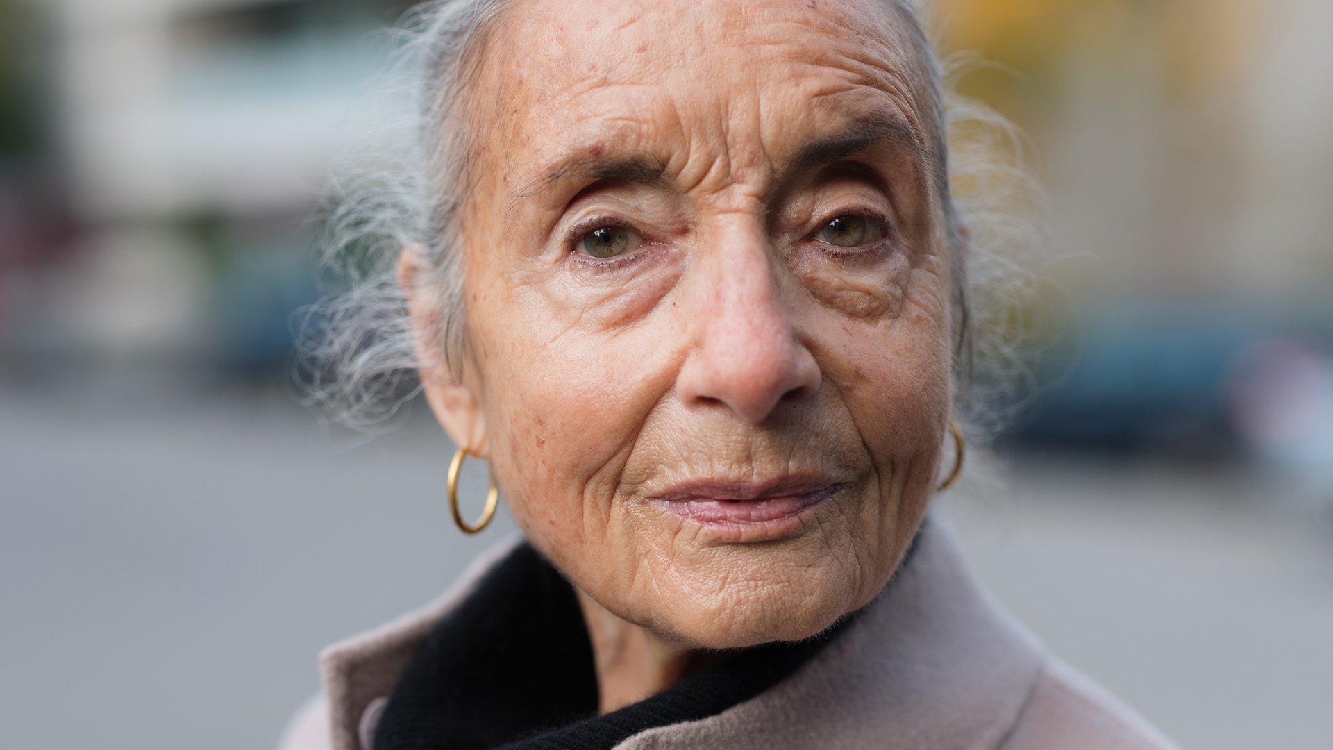 Porträt einer älteren Frau mit grauen Haaren und Ohrringen, die direkt in die Kamera blickt, unscharfer Hintergrund im Freien.
