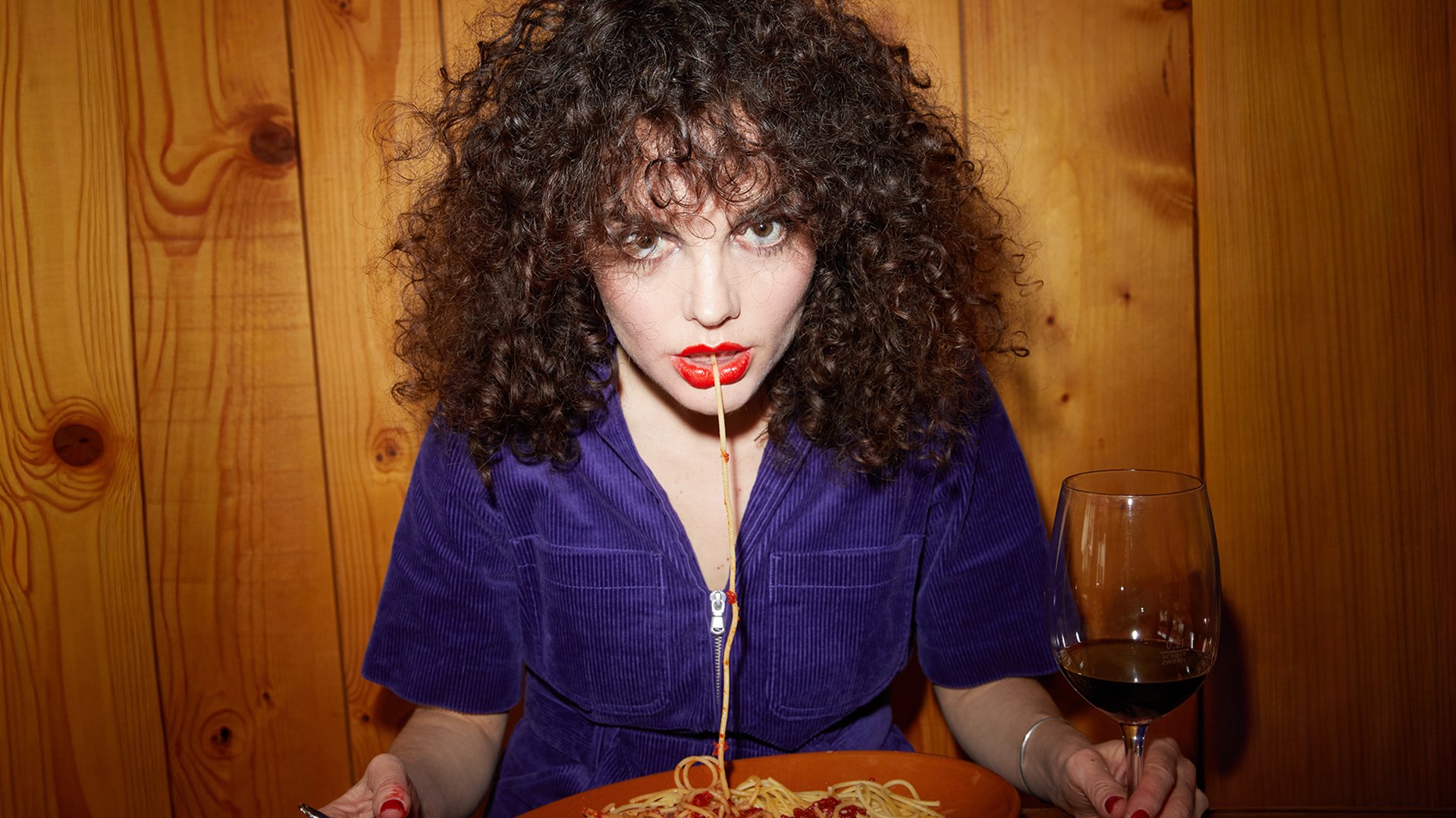 Die Künstlerin Maria de Val mit einem Teller Sgaghetti und einer Nudel im Mund und einem Weinglas in der Hand vor einer Holzvertäfelung