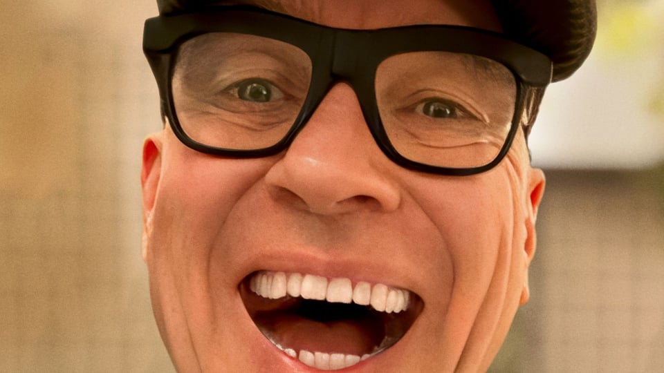Nahaufnahme eines Mannes mit schwarzer, markanter Brille. Die Person blickt direkt in die Kamera und zeigt ein breites Lachen mit sichtbaren Zähnen vor unscharfem hellem Hintergrund.