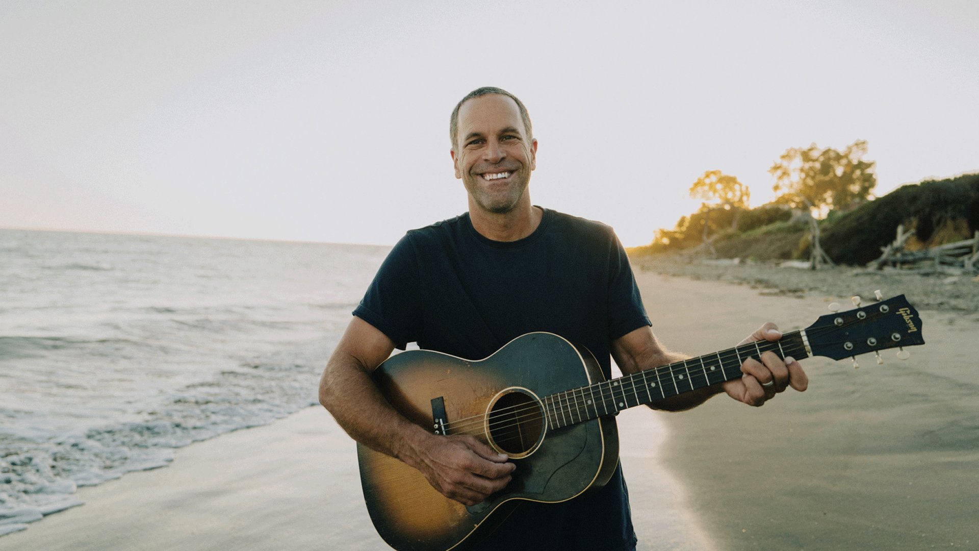 Jack Johnson mit Akustikgitarre steht bei Sonnenuntergang am Strand. Im Hintergrund sind das Meer, die Brandung und eine Küstenlandschaft mit Bäumen zu sehen.