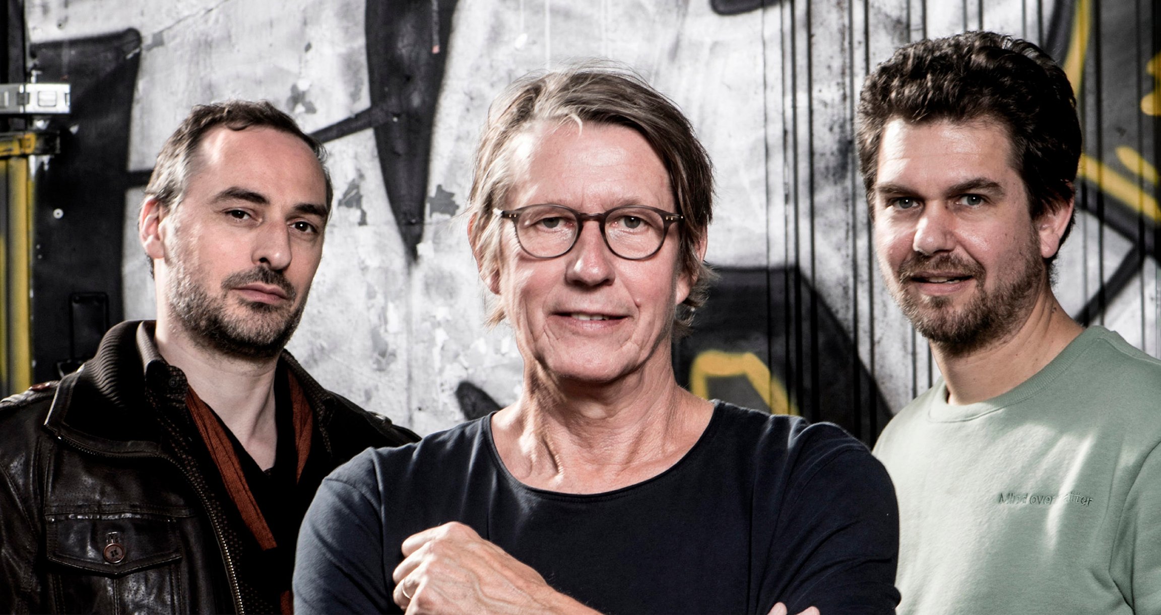 Foto von Andreas Rebers, Samuel Halscheidt (Gitarre) und André Matov vor einer Wand mit Graffiti.