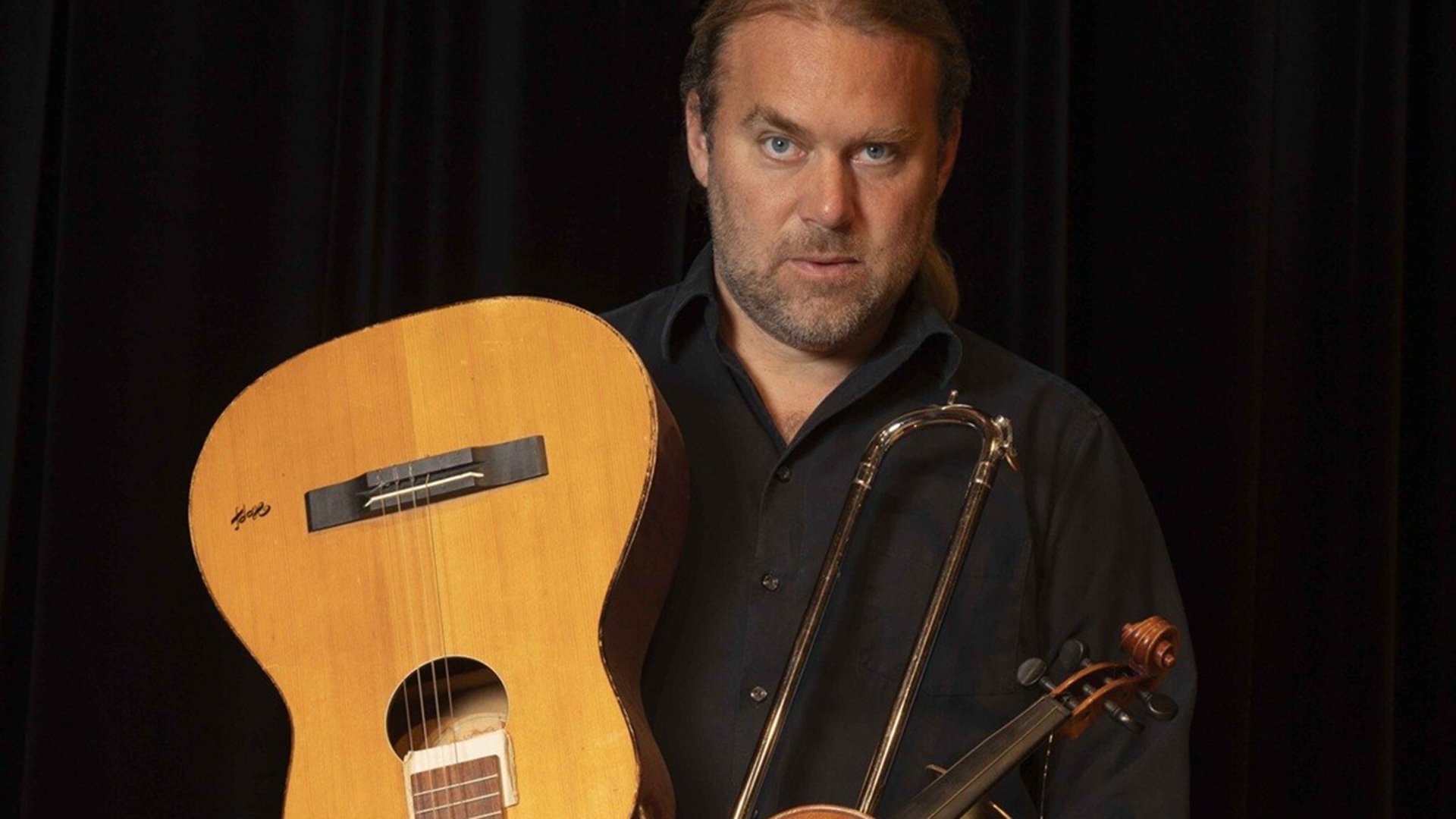 Andreas Martin Hofmeier in einem dunklen Hemd mit Gitarre und weiteren Instrumenten vor einem dunklen Vorhang