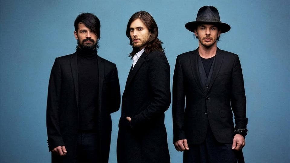 Drei Mitglieder der Band **Thirty Seconds To Mars** stehen nebeneinander vor einem blauen Hintergrund, gekleidet in schwarze, elegante Outfits; einer trägt einen Hut, die anderen haben längeres Haar und einen ernsten Blick in die Kamera.