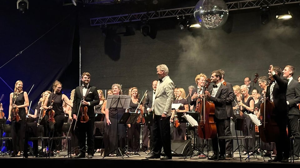 Ein Orchester mit zahlreichen Musikern mit Instrumenten auf einer Bühne, an der Decke eine große Discokugel