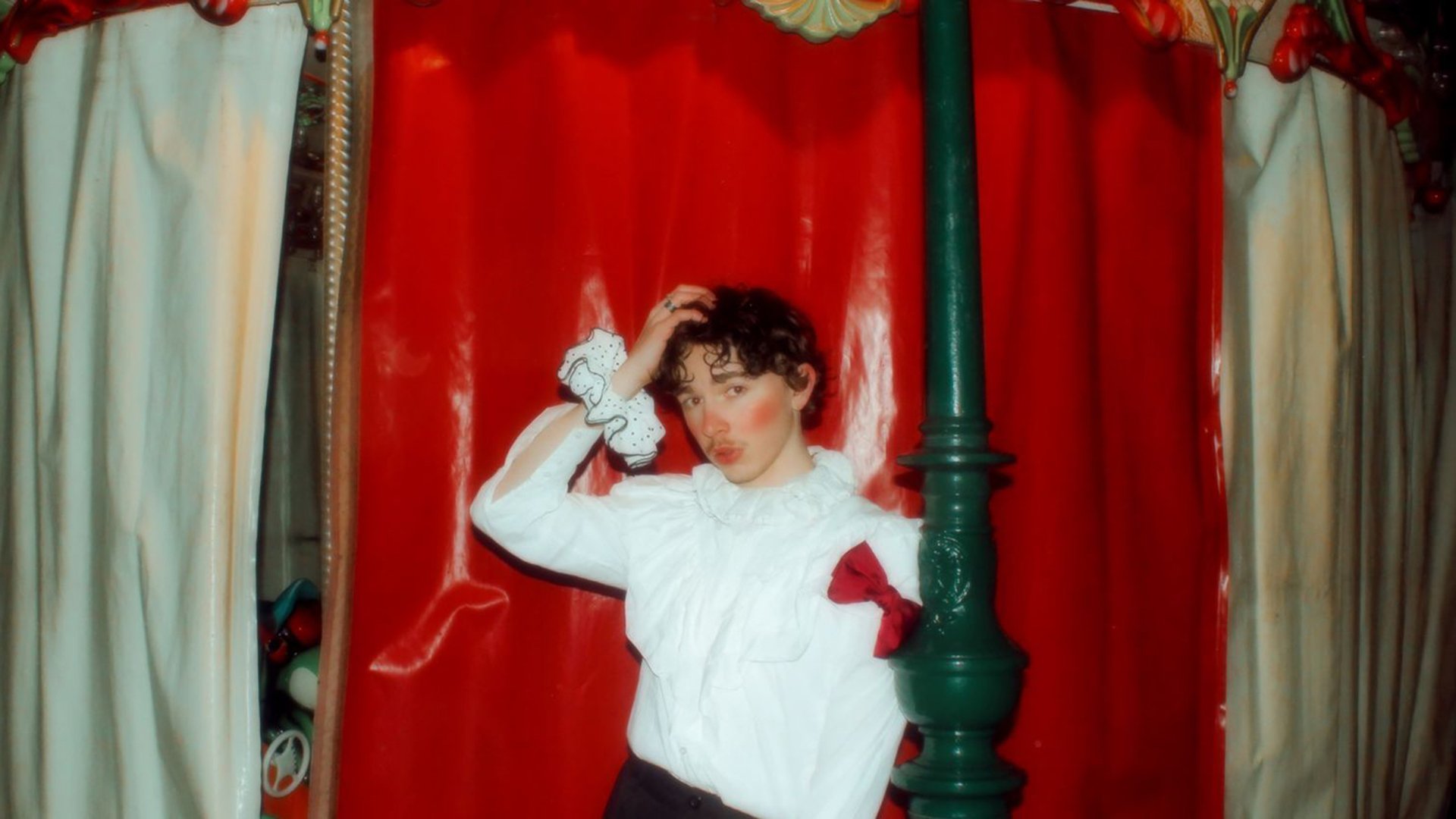 Der Künstler Oskar Haag steht vor rotem Vorhang in einer dekorativen Kulisse, mit weißer Bluse und dunkler Hose, eine Hand im Haar, daneben eine grüne Säule.