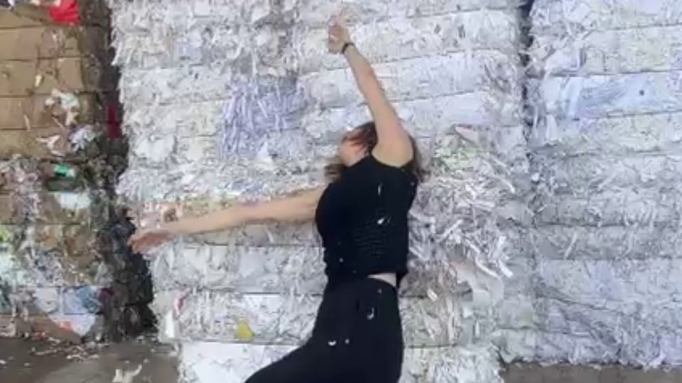 Tänzerin in schwarzer Kleidung bewegt sich vor einer strukturierten weißen Wand, umgeben von Papierfragmenten.