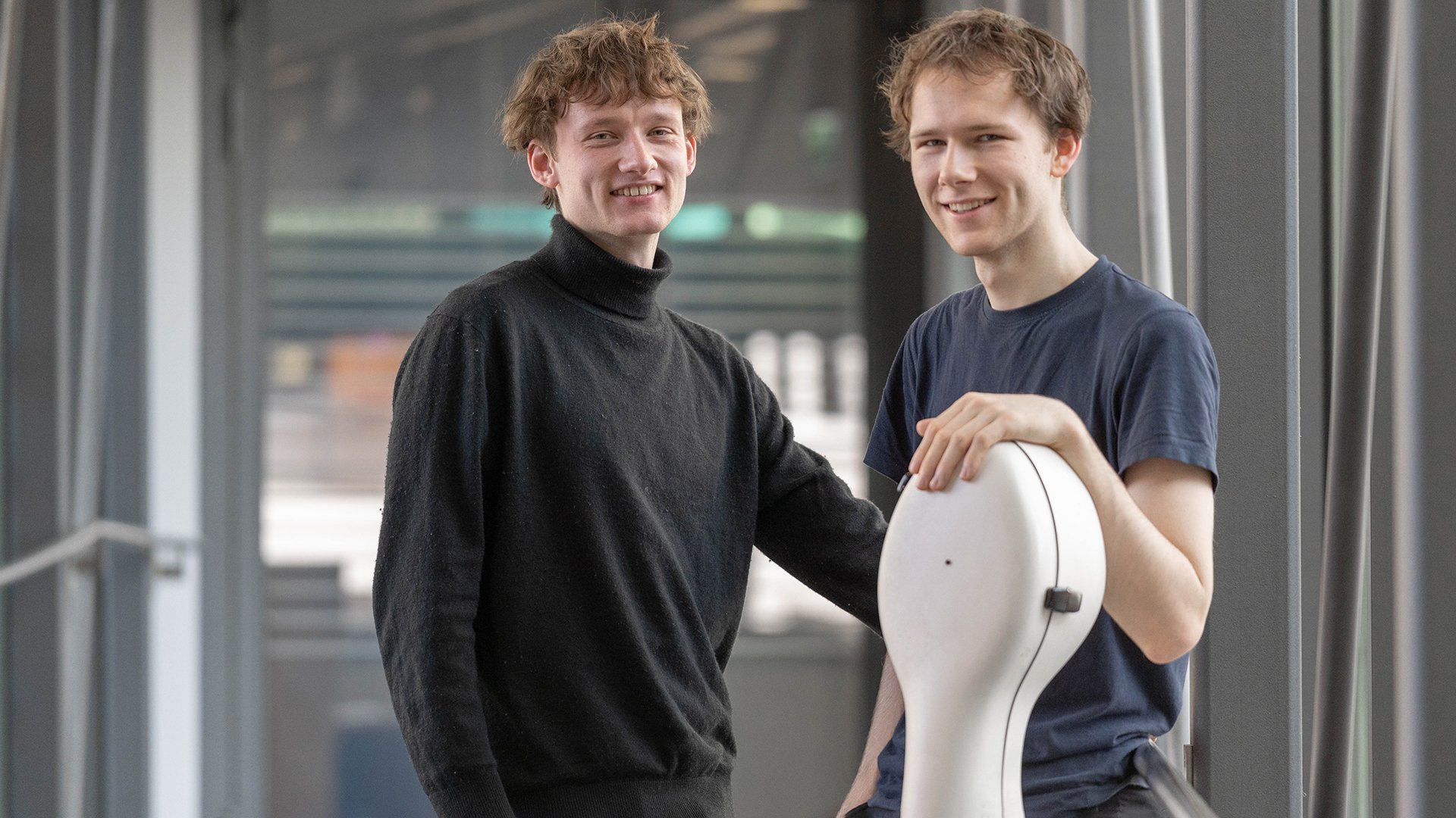 Zwei Musiker stehen in einem modernen Gebäude. Eine Person lehnt an einem Violoncello-Koffer, die andere steht daneben