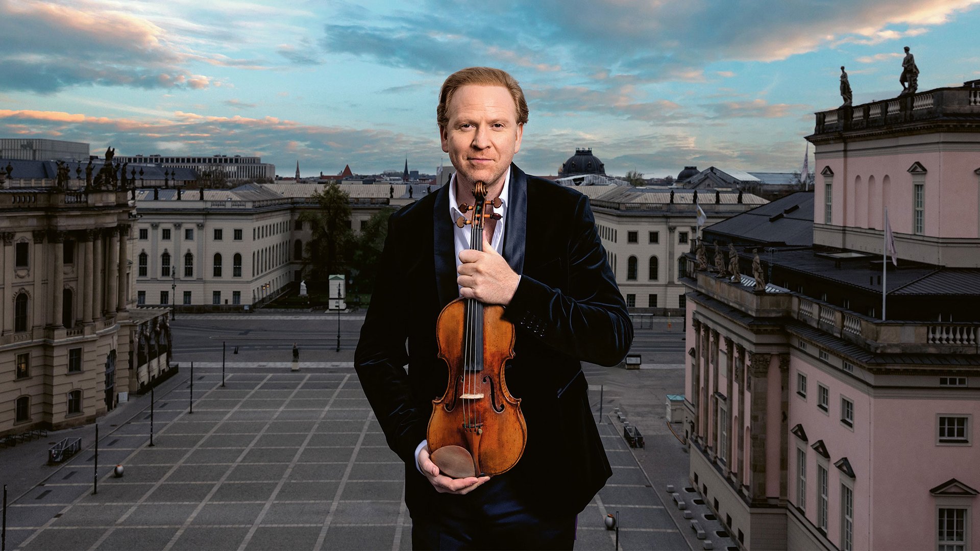 Daniel Hope in dunklem Anzug hält eine Violine vor klassischer Stadtarchitektur mit blau‑orangefarbenem Himmel und großem Stadtplatz im Hintergrund 