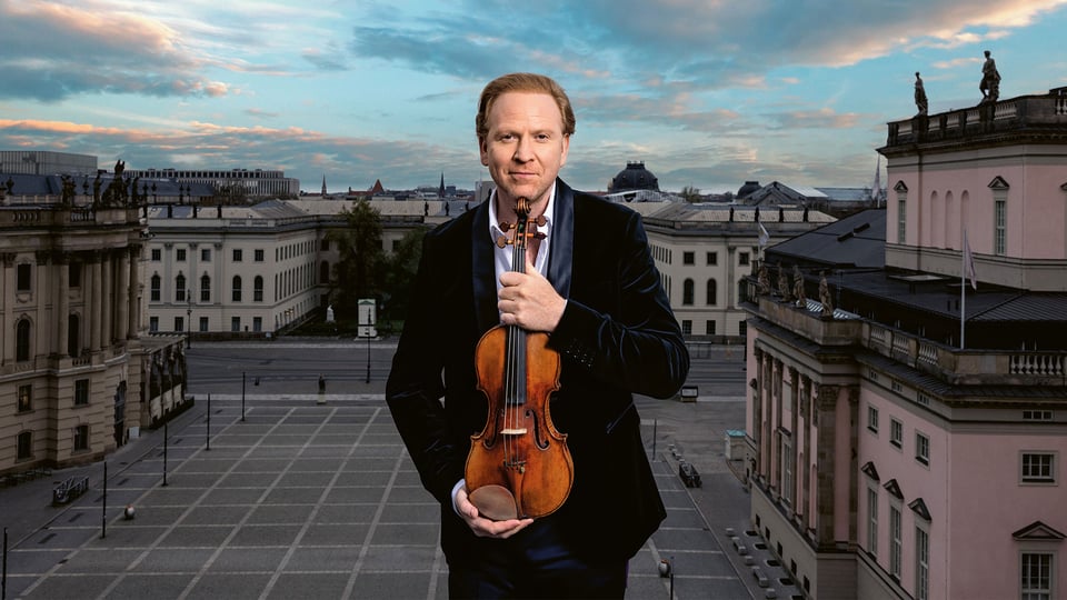 Daniel Hope in dunklem Anzug hält eine Violine vor klassischer Stadtarchitektur mit blau‑orangefarbenem Himmel und großem Stadtplatz im Hintergrund 