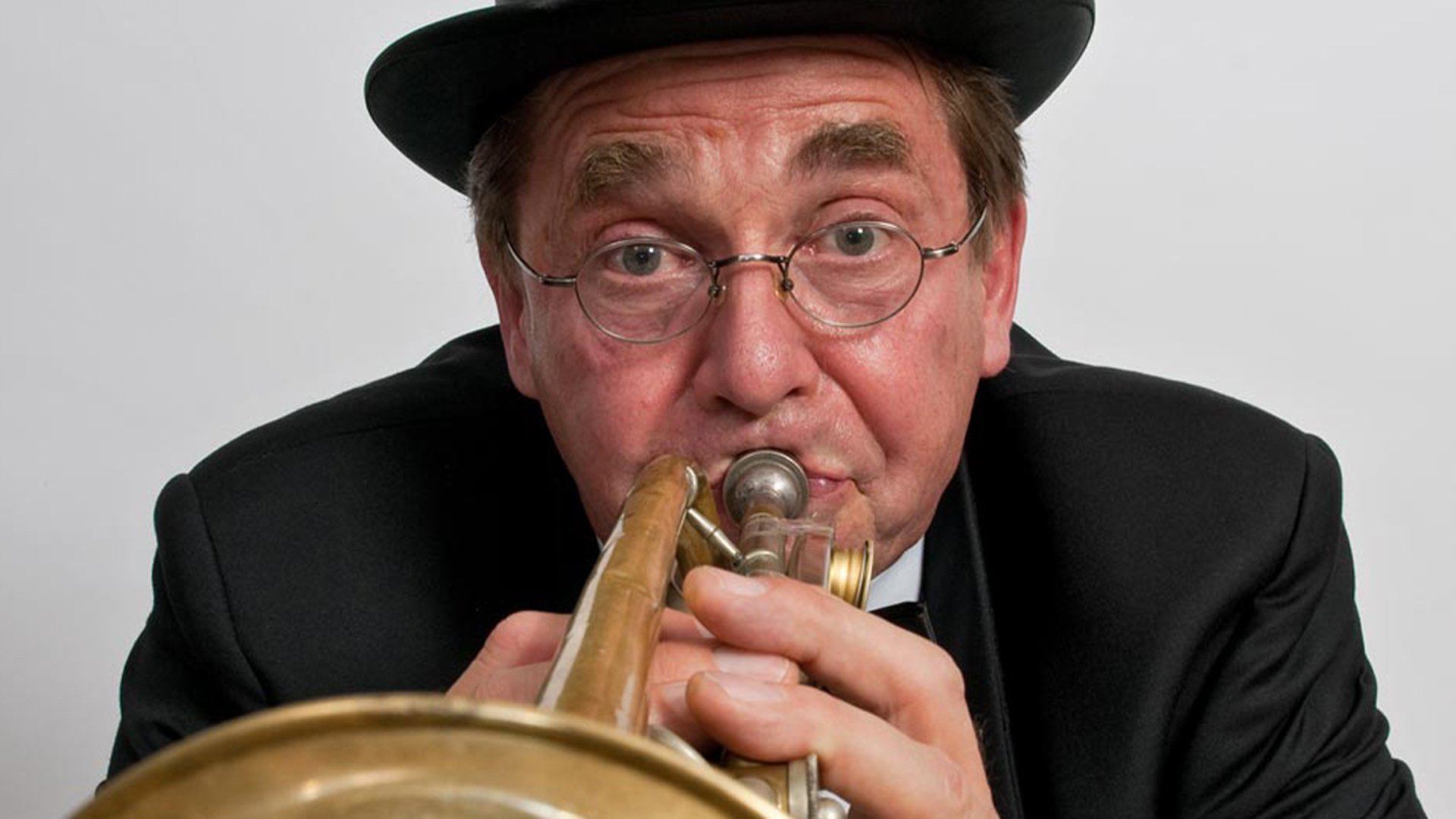 Nahaufnahme einer Person mit Hut und Brille, die eine Trompete direkt in die Kamera spielt, vor hellem Hintergrund.