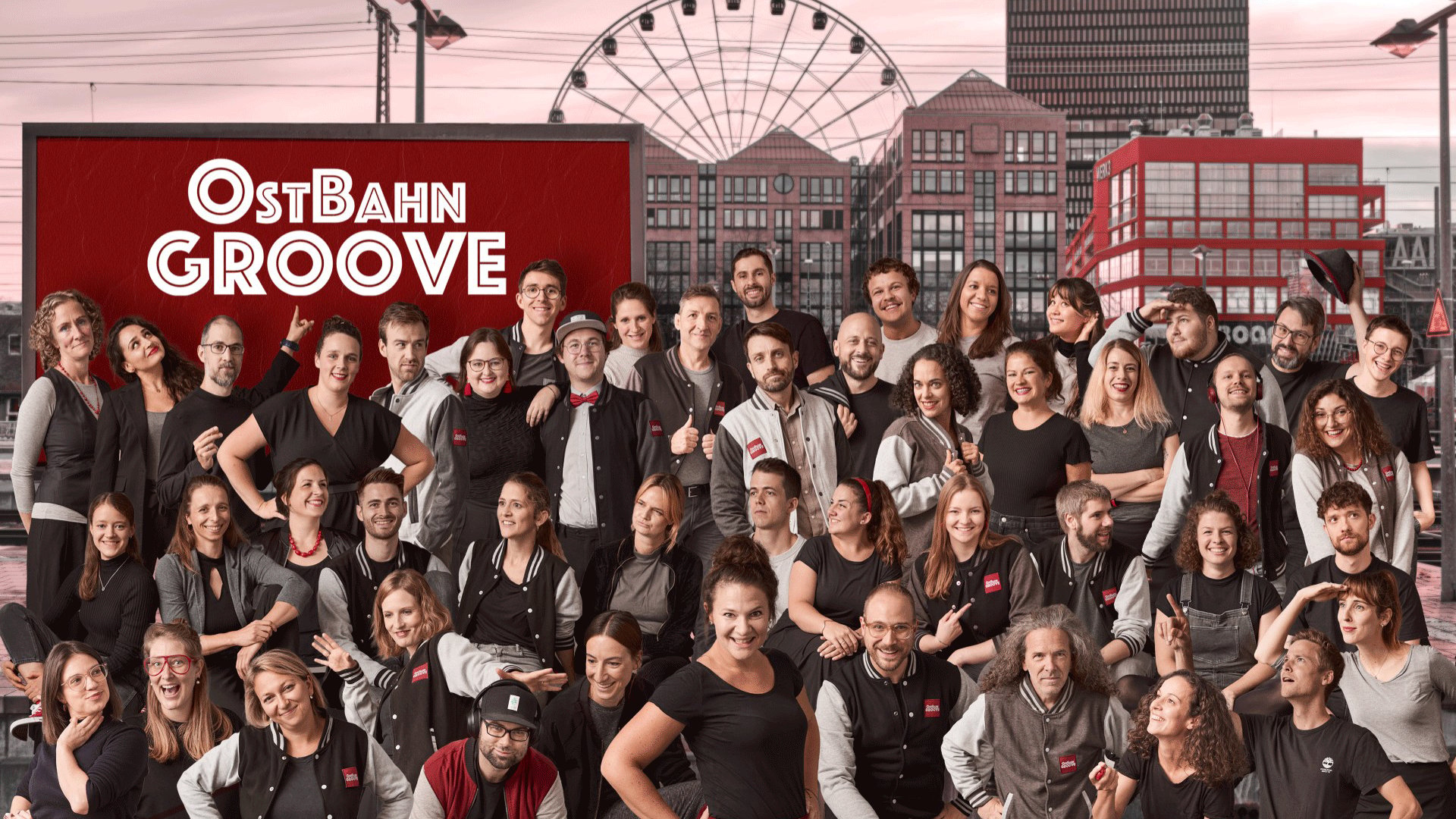 Gruppenfoto des Chors OstBahn Groove vor einer urbanen Kulisse mit Gebäuden und Riesenrad. Zahlreiche Chormitglieder in schwarz grauer Kleidung und Collegejacken sind versetzt angeordnet, während im Hintergrund ein rotes Schild mit der Aufschrift OstBahn Groove zu sehen ist.