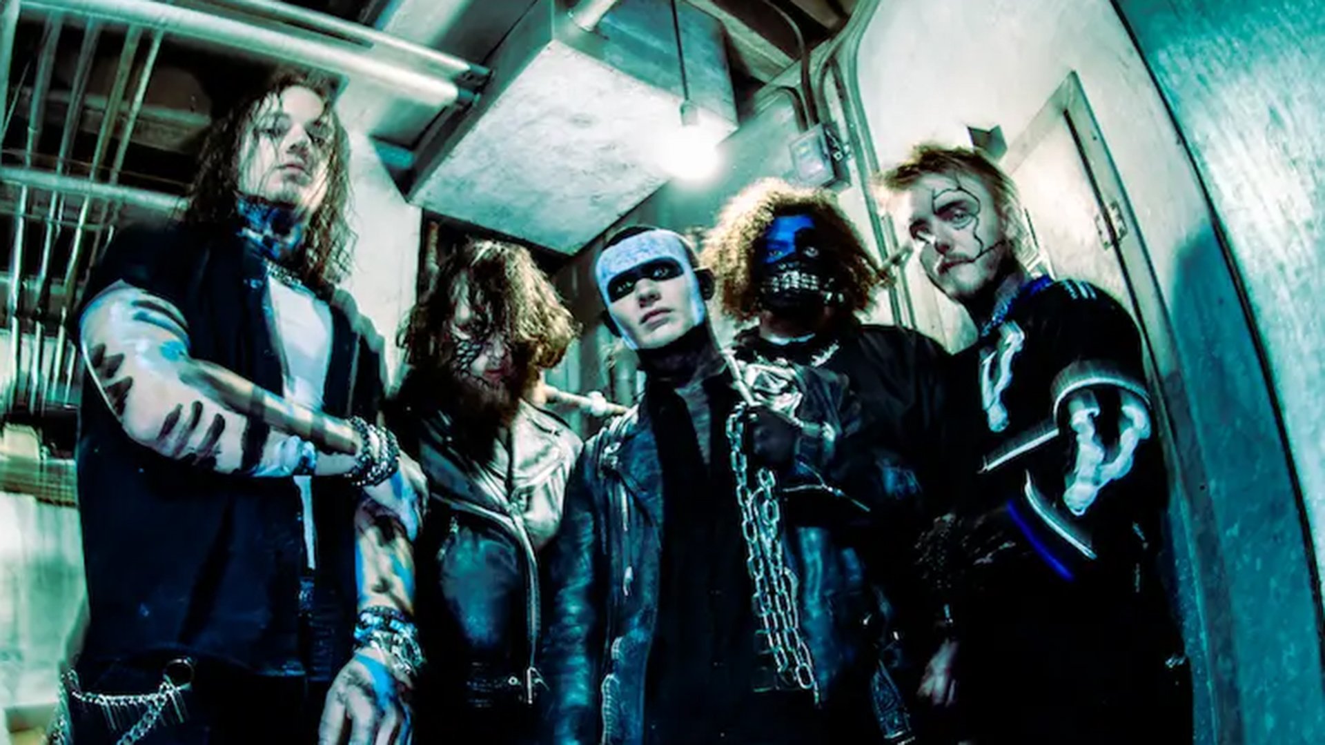 Fünf Musiker stehen eng zusammen in einem Industrieflur mit Rohren und Neonlicht, auffälliges Make-up und dunkle Kleidung, düstere Metal-Ästhetik