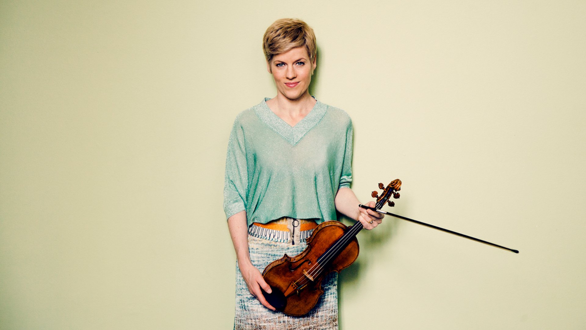 Isabelle Faus in türkisem Pullover mit Streichinstrument blickt lächelnd in die Kamera, vor pastellgrünem Hintergrund
