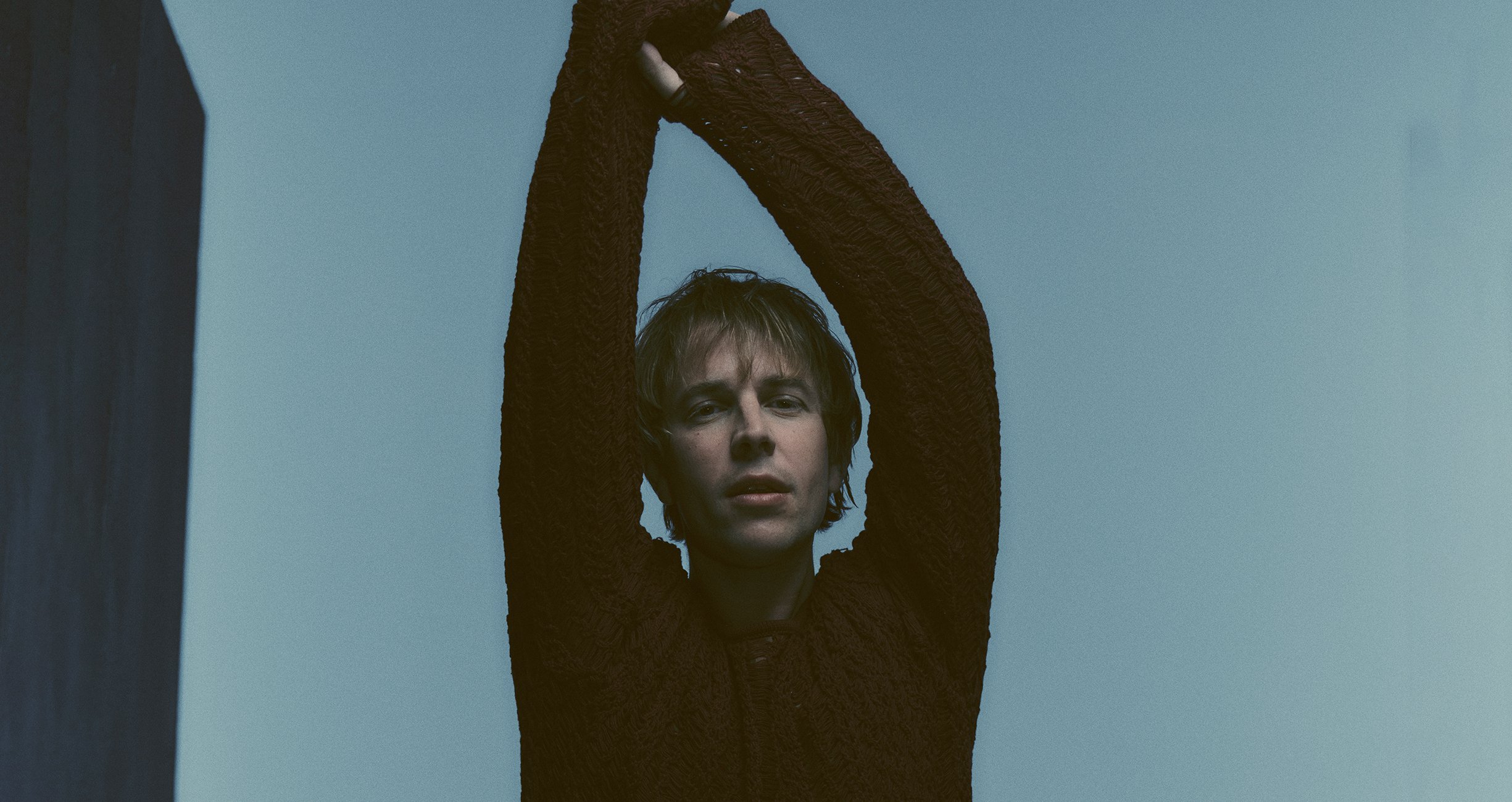 Tom Odell steht vor einem blauen Hintergrund und hält seine Arme nach oben