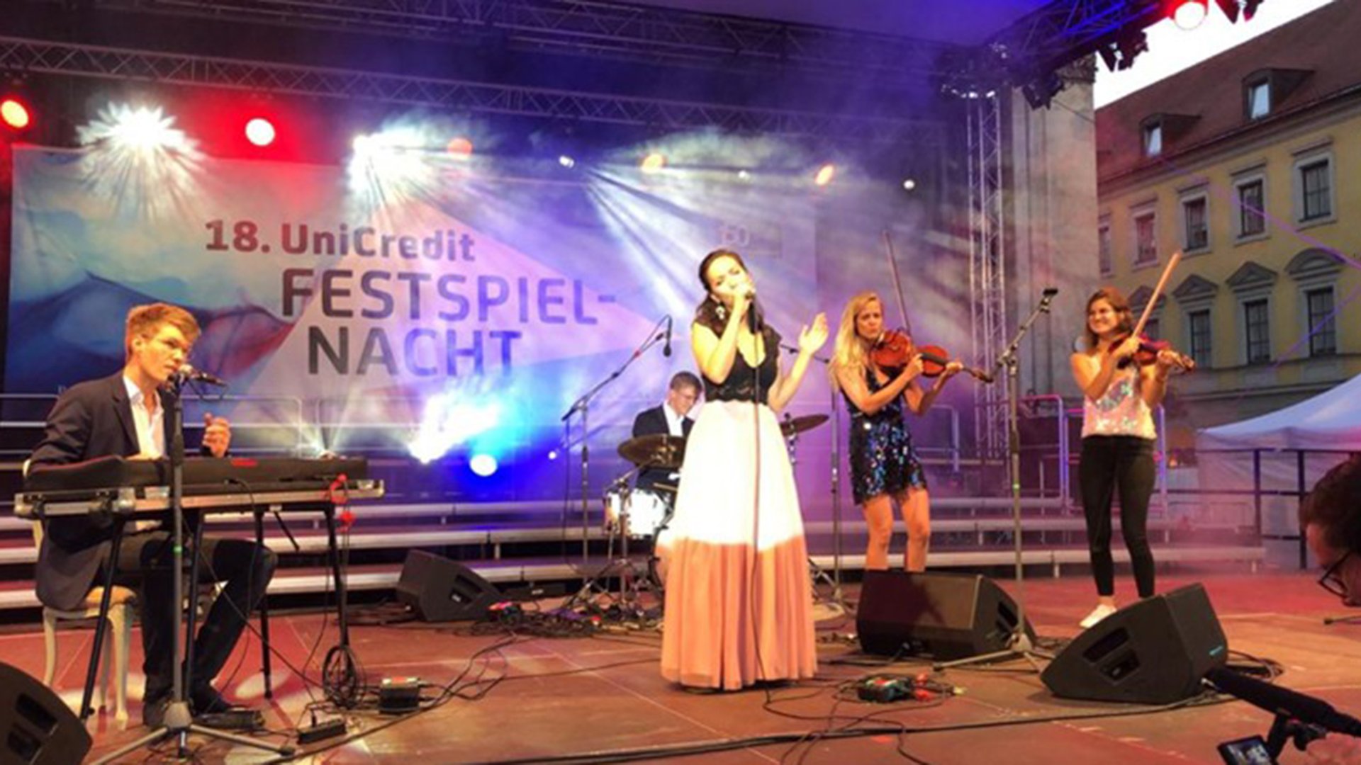 Veranstaltungsfoto eines Open‑Air‑Konzerts: Mehrere Musikerinnen und Musiker stehen auf einer beleuchteten Bühne im Außenbereich. In der Mitte singt eine Person in ein Mikrofon, begleitet von Keyboard, Schlagzeug und zwei Geigen. Farbiges Bühnenlicht und Nebeleffekte sind sichtbar. Im Hintergrund hängt ein Banner mit der Aufschrift „18. UniCredit FESTSPIEL‑NACHT“. Das Bild zeigt eine Live‑Konzertatmosphäre mit Publikum und historischer Gebäudeumgebung.