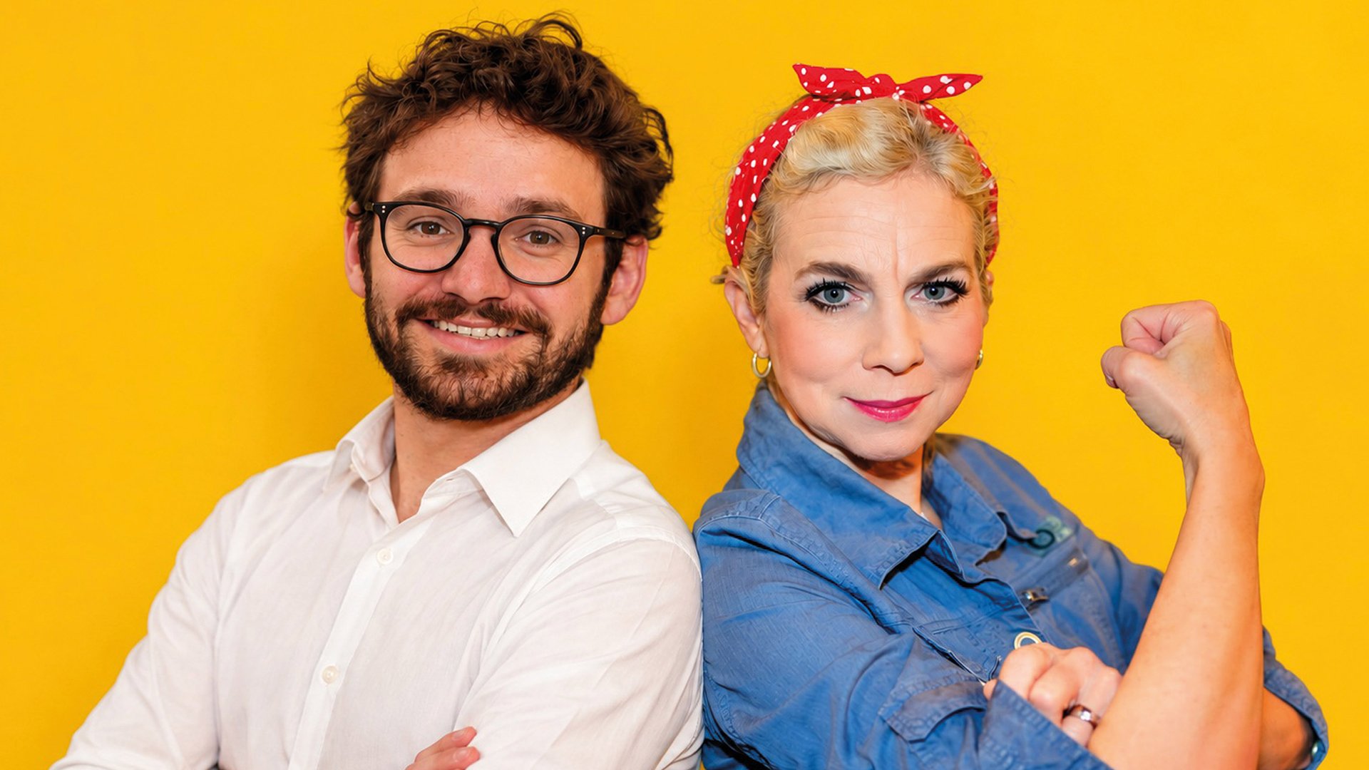 Franziska Ball und Felix Mathy stehen aneinandergelehnt, sie mit rot getupftem Kuopftuch und eine Hand zur Faust geballt, er in weißem Hemd vor gelbem Hintergrund 