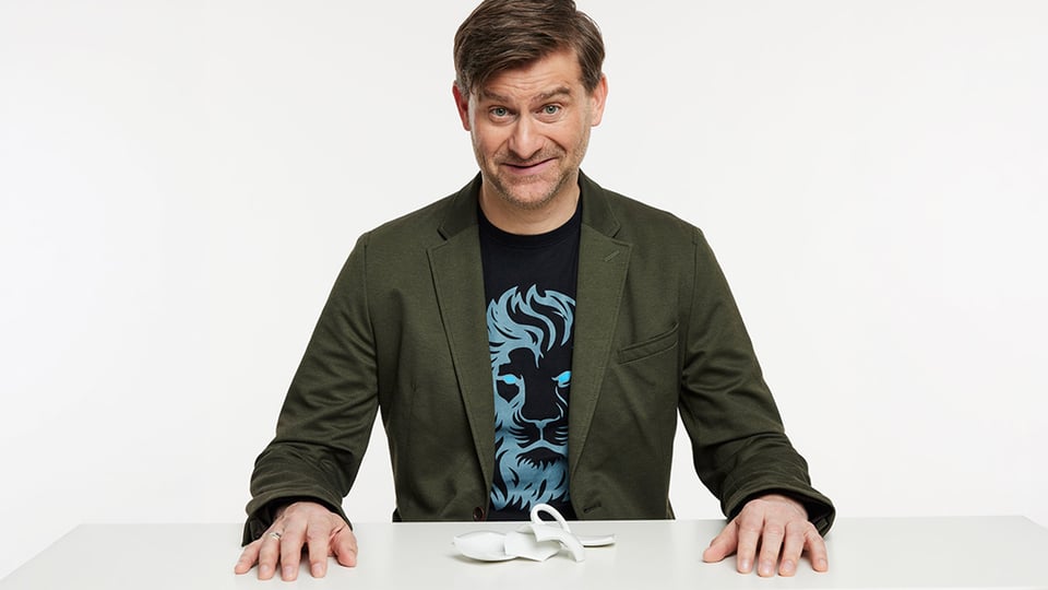 Michael Altinger an einem weißen Tisch vor hellem Hintergrund, in grünem Sakko und dunklem T Shirt mit Löwenmotiv, vor sich eine zerbrochene weiße Tasse auf der Tischplatte.