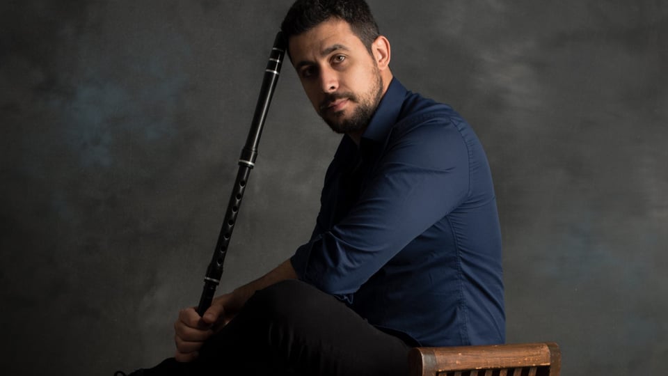 Foto vom Event Zhivko Vasilev & Harmelogic feat. Rayna Vasileva mit dem Künstler, der auf einem Stuhl sitzt und ein Blasinstrument in der Hand hält, vor grauem Hintergrund.