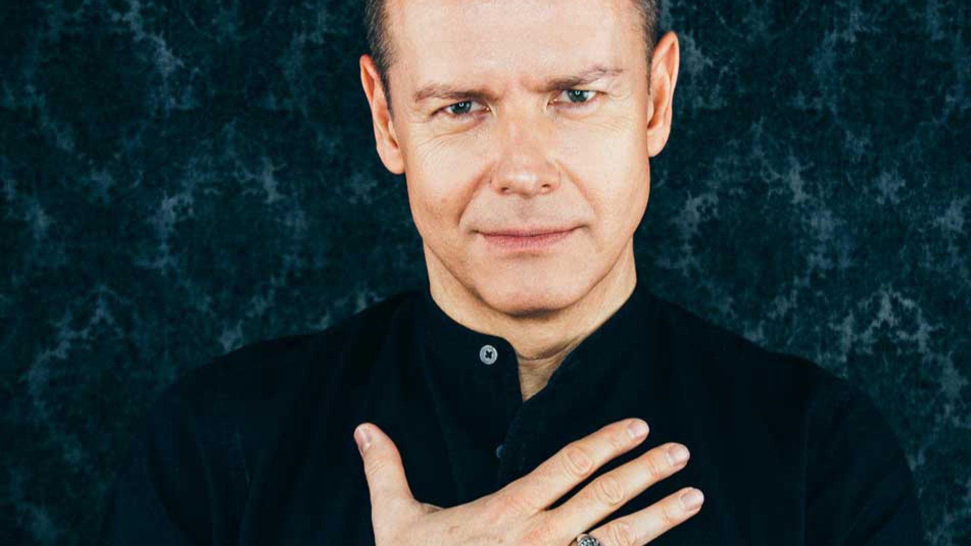 Ein Foto für das Event von Lars Ruth mit dem Künstler, der eine Hand auf der Brust hat, vor einem Hintergrund mit einem Muster