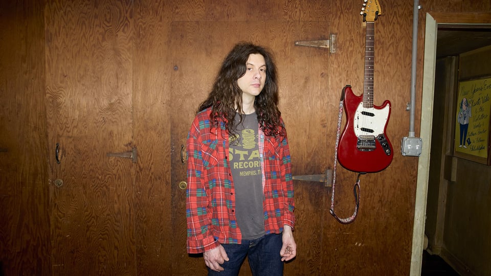 Kurt Vile steht vor einer Holzwand neben einer roten E-Gitarre, trägt ein kariertes Hemd und ein T-Shirt, direkter Blick in die Kamera.
