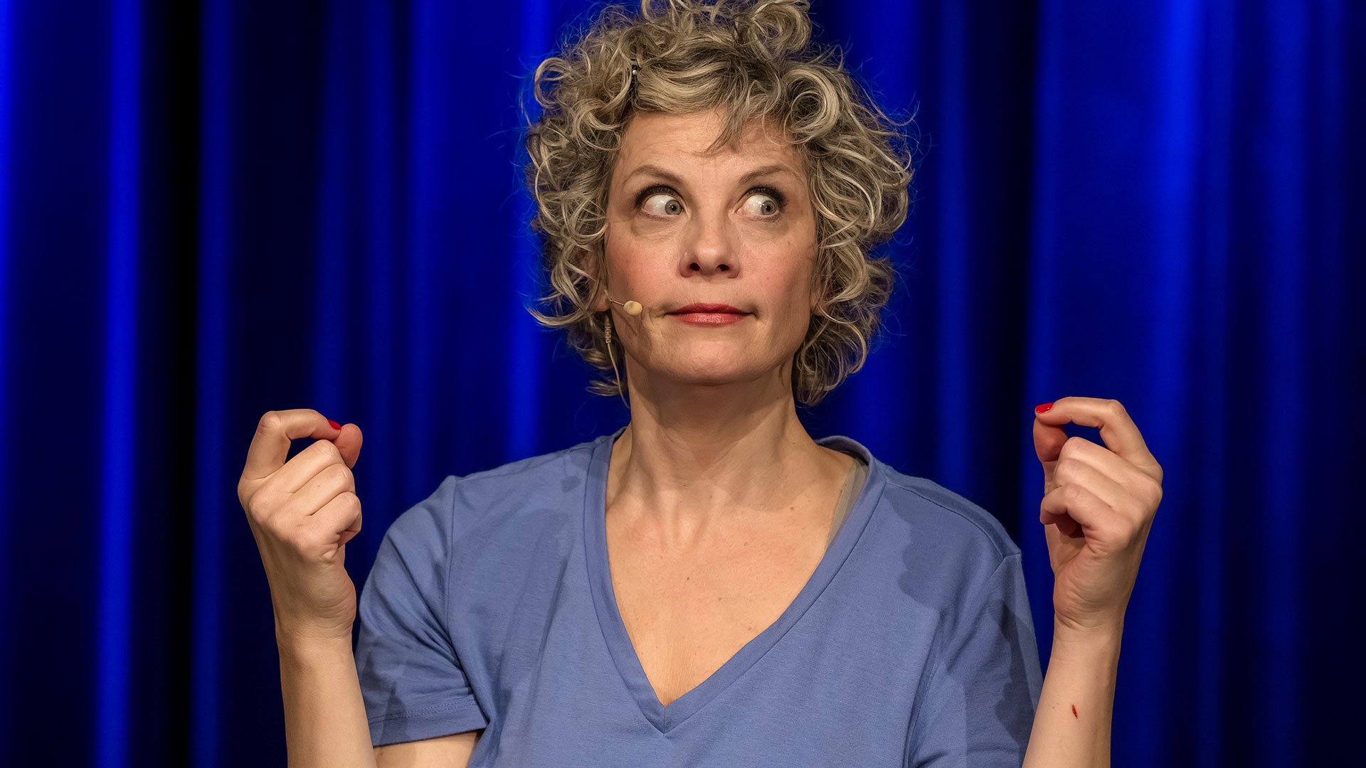 Pressefoto zu „Anna Schäfer – Schlafende Hunde – Ein hochkomischer Nachtflug“ mit Anna Schäfer auf einer Bühne vor blauem Hintergrund, mit Mikrofon am Gesicht und erhobenen Händen.