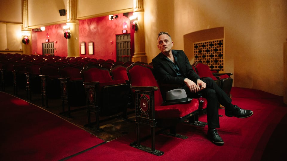 Eventmotiv zu Dave Hause & The Mermaid: Person sitzt allein in einem historischen Theatersaal mit roten Polstersitzen und rotem Teppich, trägt dunkle Kleidung; auf dem Nachbarsitz liegt ein Hut. Warme Innenbeleuchtung, ruhige Konzertatmosphäre vor einer Live‑Show.