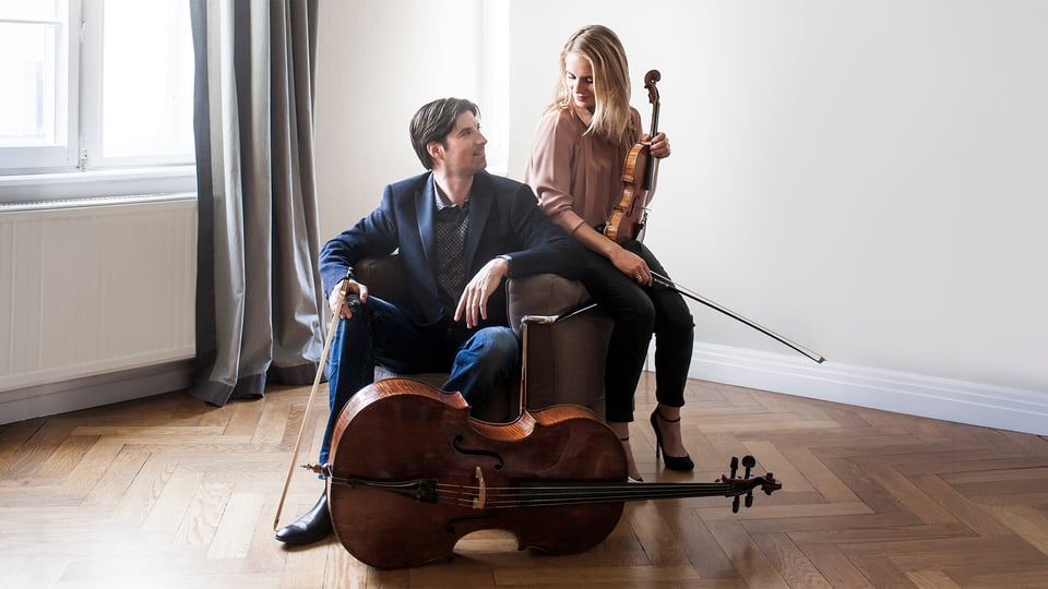 Zwei Musiker mit Violoncello und Violine sitzen in einem hellen Raum mit Fenster und Vorhang; das Cello liegt im Vordergrund auf dem Boden.