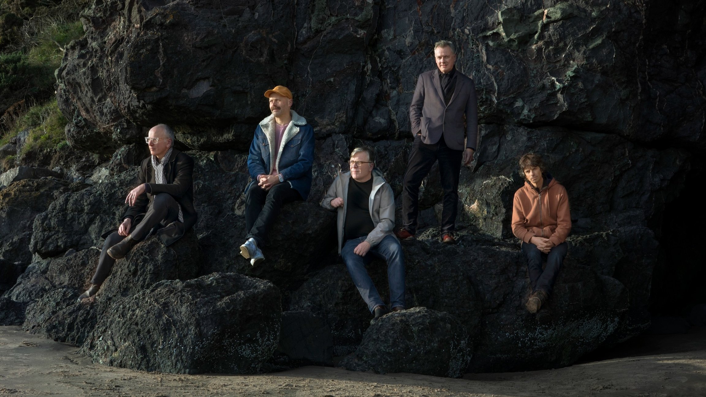Die Band Teenage Fanclub sitzen und stehen auf dunklen Felsen an einer Küste, im Vordergrund Sandstrand, im Hintergrund eine steile Felswand.
