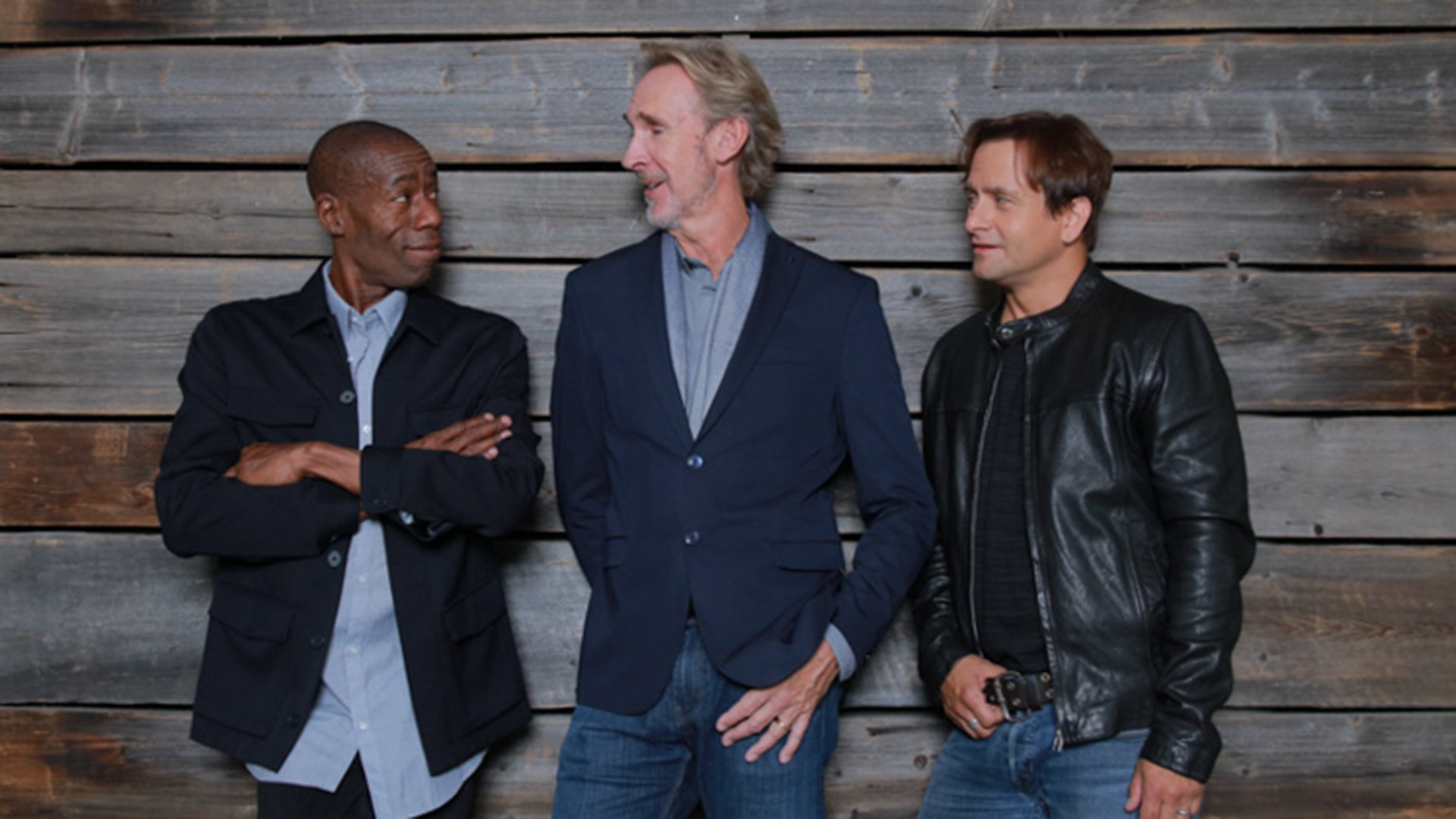 Pressefoto von Mike + The Mechanics mit drei Bandmitgliedern in dunkler Kleidung vor einer Holzwand, darunter eine Person in Lederjacke, eine im blauen Sakko und eine mit verschränkten Armen
