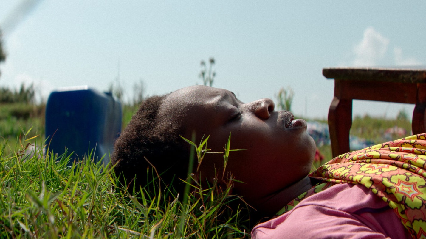 Filmszene aus Imana mit einer Person im rosa Oberteil, die im Gras liegt, daneben ein blauer Kanister und ein Holztisch, vor ländlicher Landschaft unter hellem Himmel.
