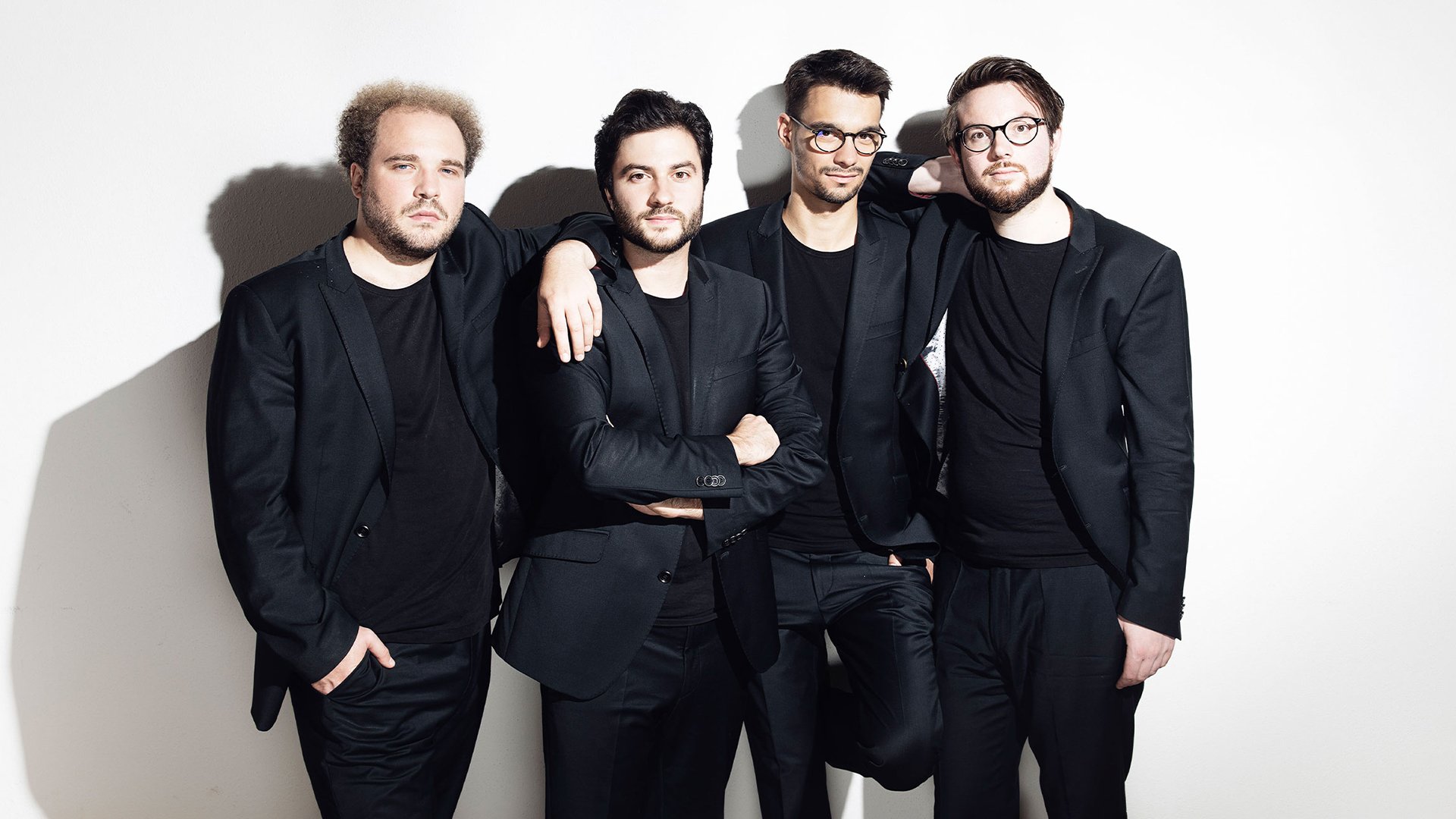 Goldmund Quartett mit vier Musiker:innen in schwarzen Anzügen vor hellem Hintergrund, nebeneinander stehend, teilweise mit verschränkten Armen oder aufgelegten Händen, ohne Instrumente im Bild sichtbar.